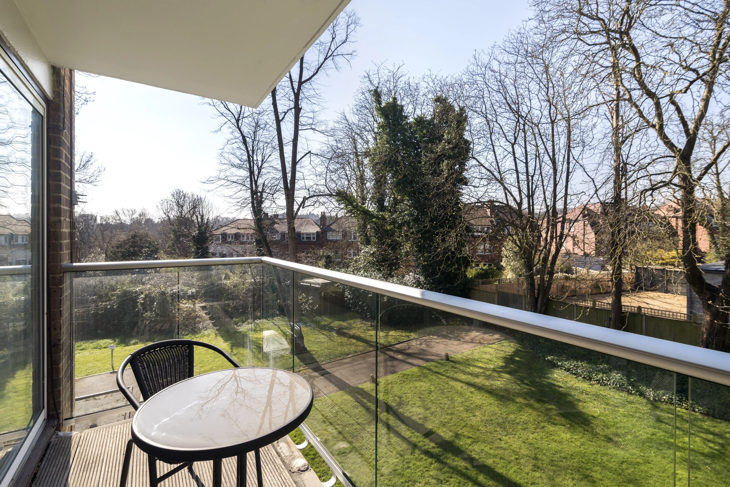 View from a balcony showing a glass railing, a round table with two chairs, a grassy yard, leafless trees, and neighboring houses under a clear blue sky.