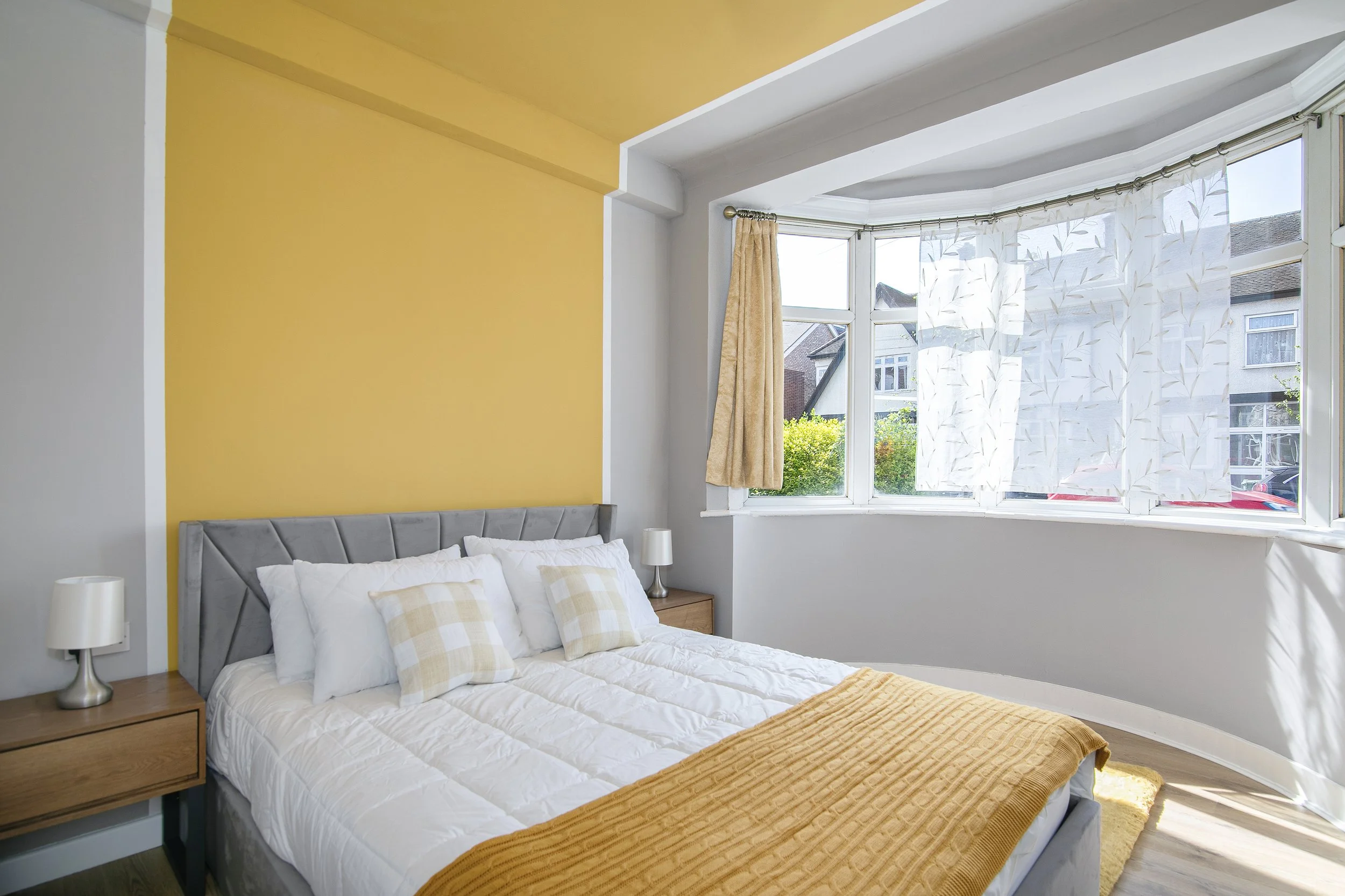 A bedroom with a bed featuring white and beige checkered pillows, a mustard yellow bedspread, and a gray upholstered headboard. There are two white bedside lamps on wooden nightstands. The room has a large bay window with white curtains decorated wit