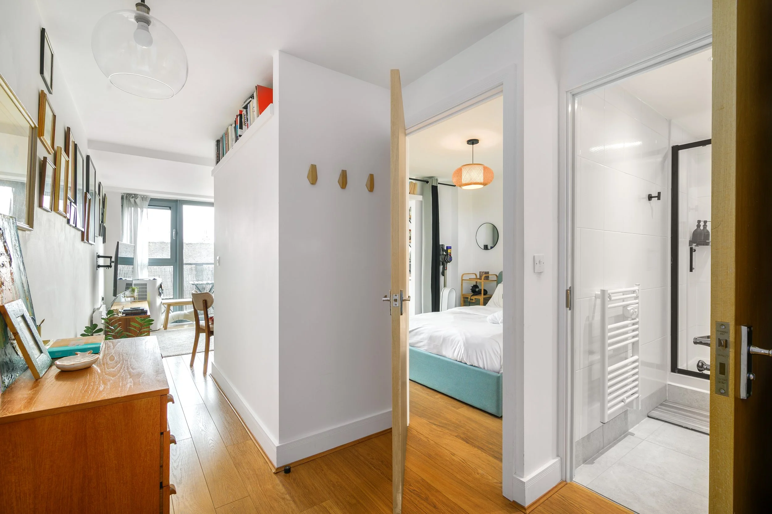 A view of an apartment interior showing a living room and bedroom area, with a partial view of a bathroom. The living room has a gallery wall with framed pictures, a wooden table with art supplies, and a window with sheer curtains. The bedroom has a 