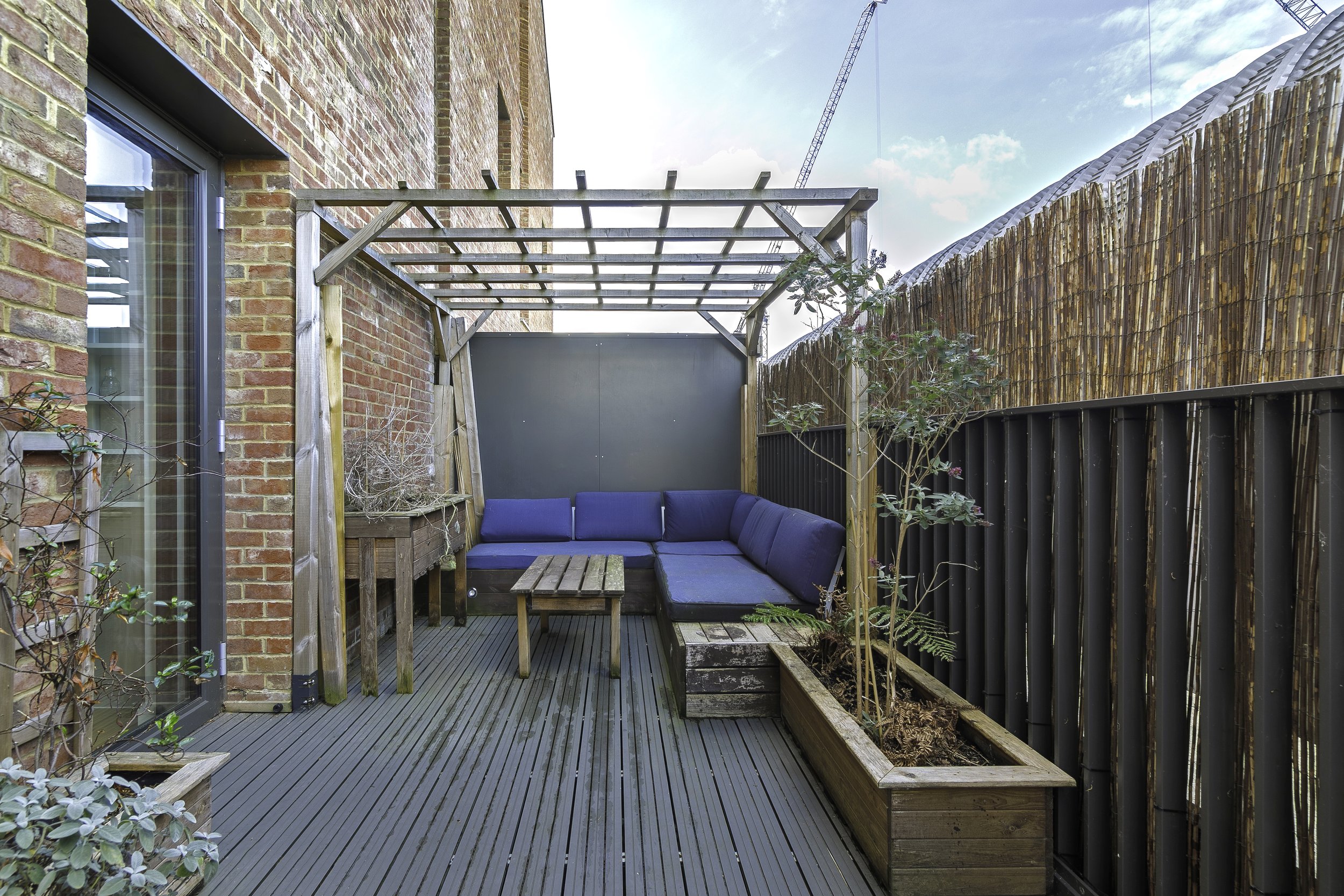 Small outdoor patio with a wooden trellis, a blue sectional sofa, and planters along a wooden fence.