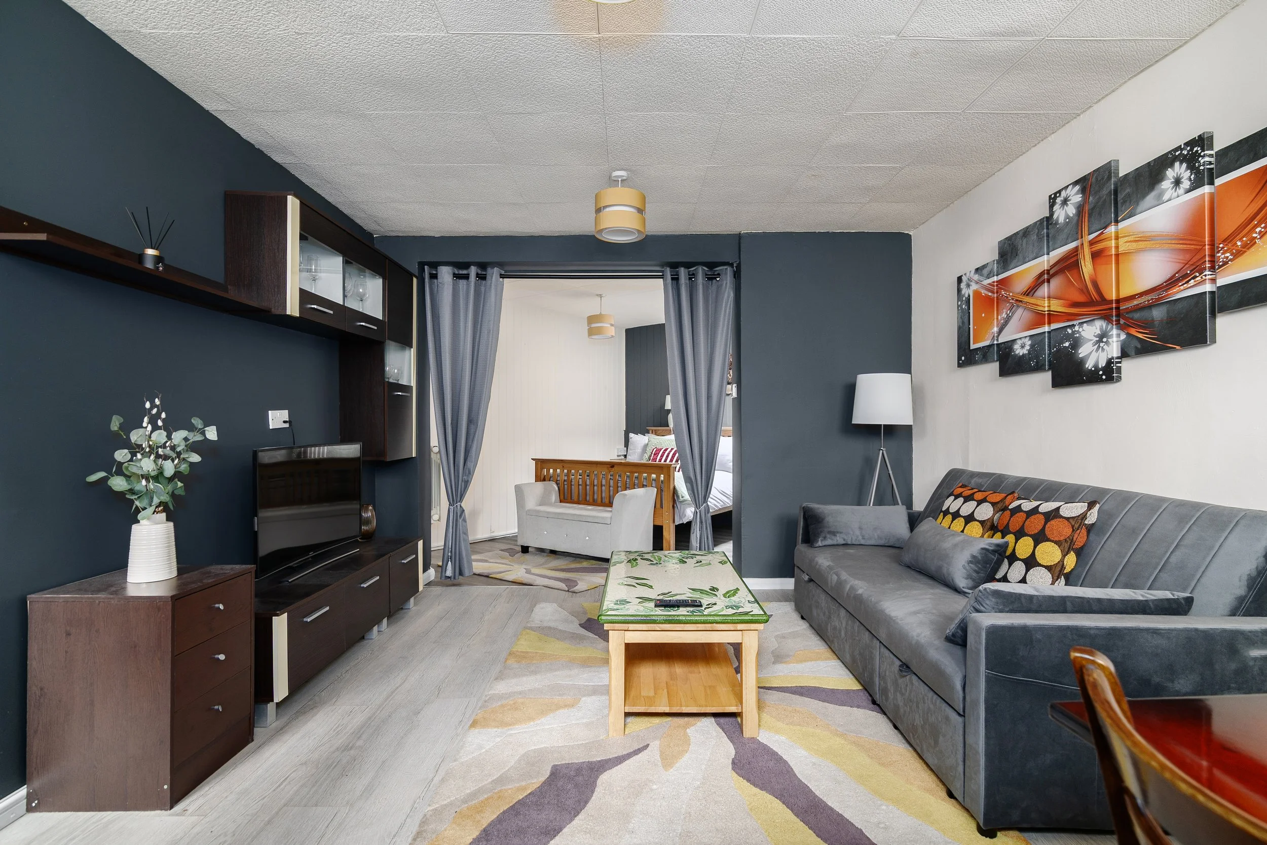 A living room with a dark blue accent wall, a gray sofa with polka dot pillows, a wooden coffee table, a large painting with orange and white swirl patterns, a television, a white vase with eucalyptus leaves, and a navy curtain separating the living 