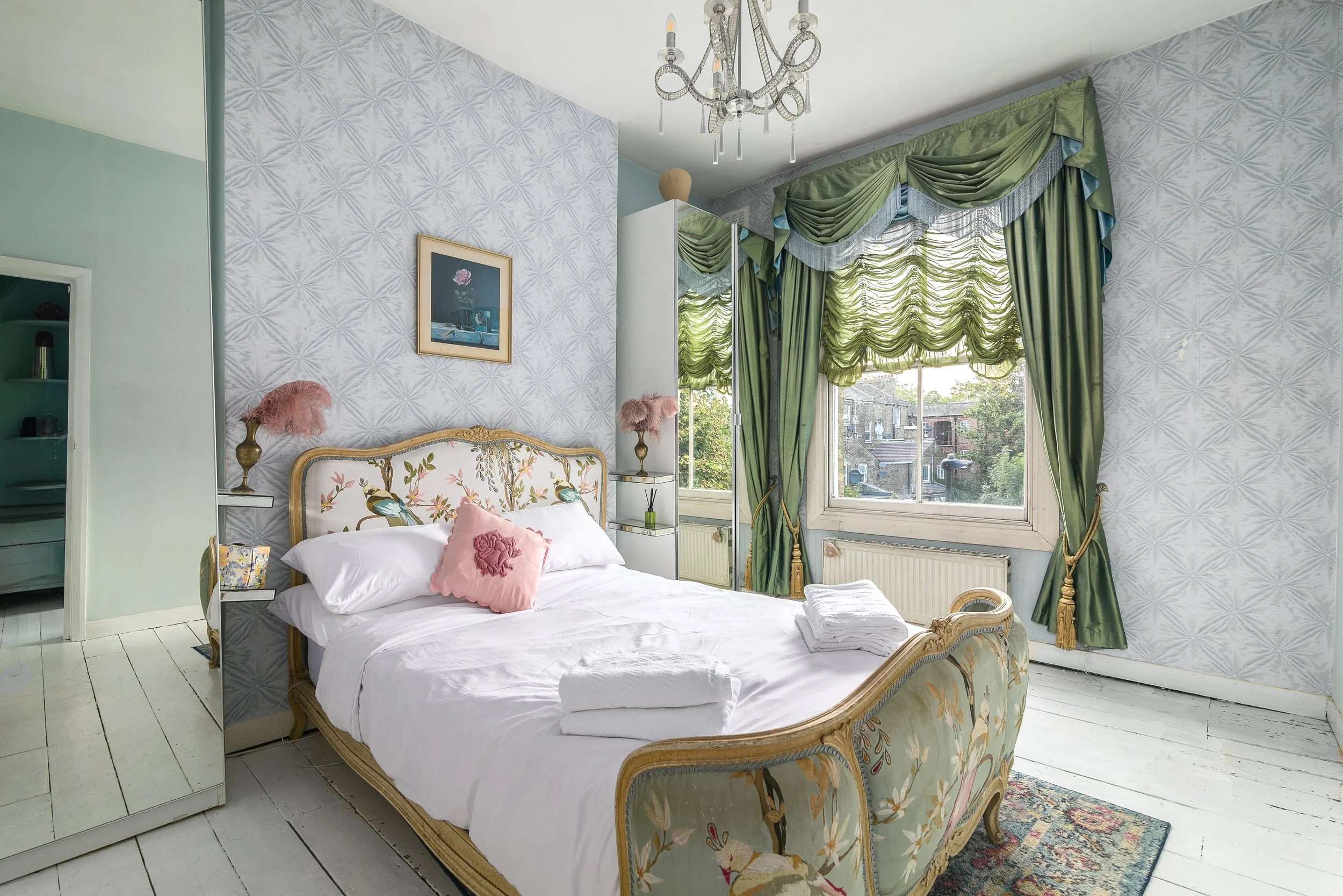 A bedroom with a vintage upholstered bed, pink decorative pillows, and a white bedspread. The room has light-colored wooden floors, patterned wallpaper, and large windows with elaborate green draped curtains. There is a chandelier hanging from the ce