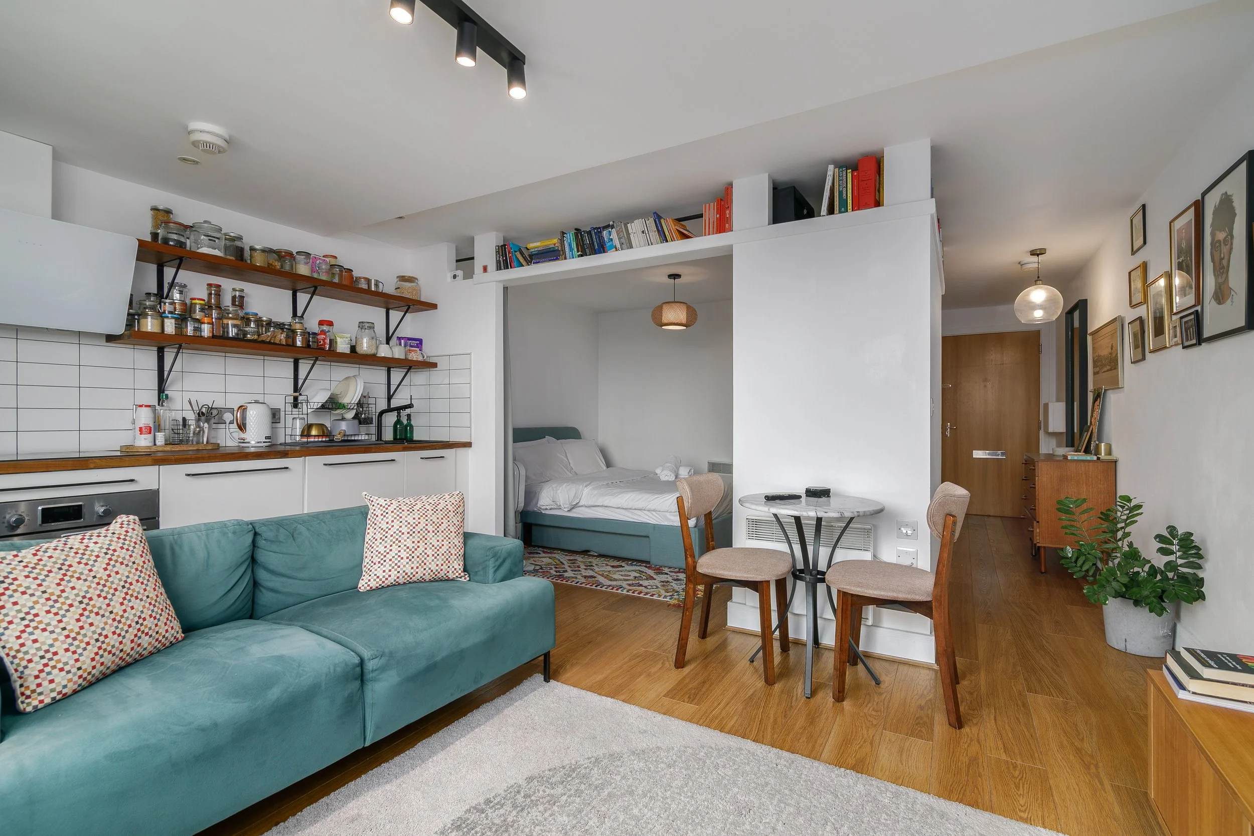Interior of a modern apartment with a green sofa, a small round table with chairs, a kitchen area with open shelves, a bed in a nook, and wall art including a Frida Kahlo poster.