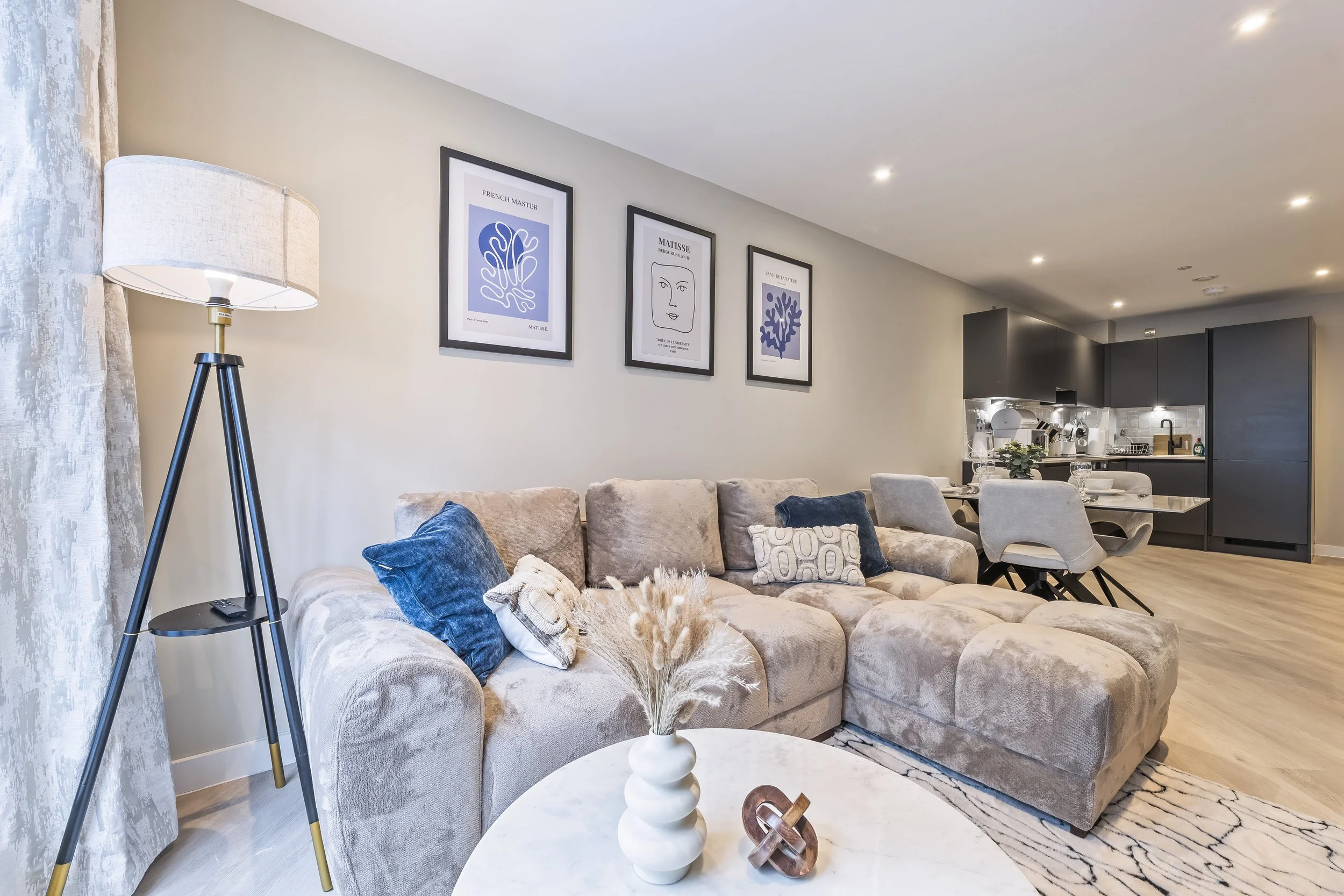 Living room with beige sectional sofa with cushions, a coffee table with decorative items, a tall tripod floor lamp, wall art featuring framed line art and abstract pieces, and a visible kitchen area with dark cabinetry and a dining table set for mea