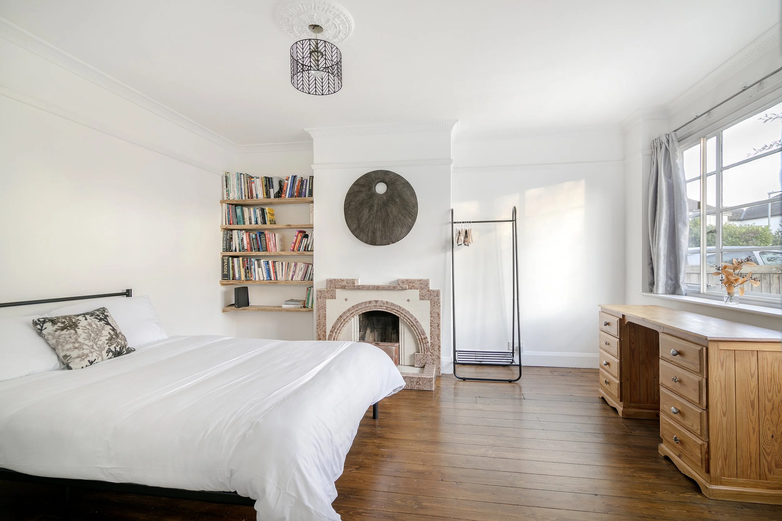 A bedroom with a white bed, a window with curtains, a bookshelf with books, a decorative wall art, a fireplace, and a wooden dresser.
