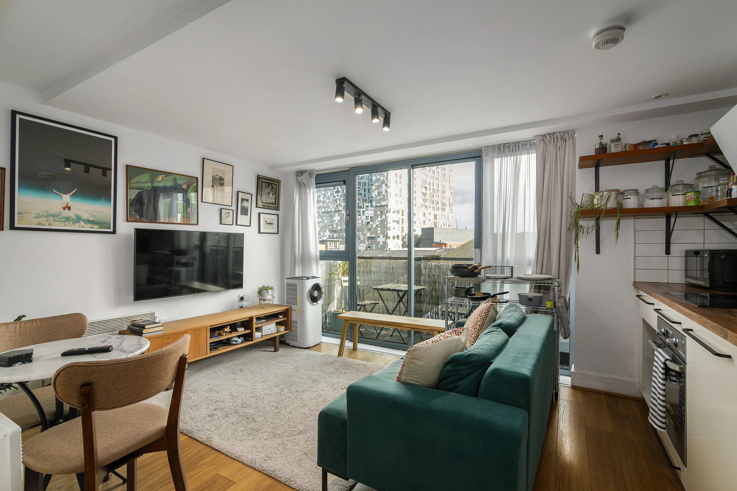 Living room with a sliding glass door leading to a balcony, decorated with framed artwork, a green sofa, a dining table with chairs, and kitchen shelves with jars.