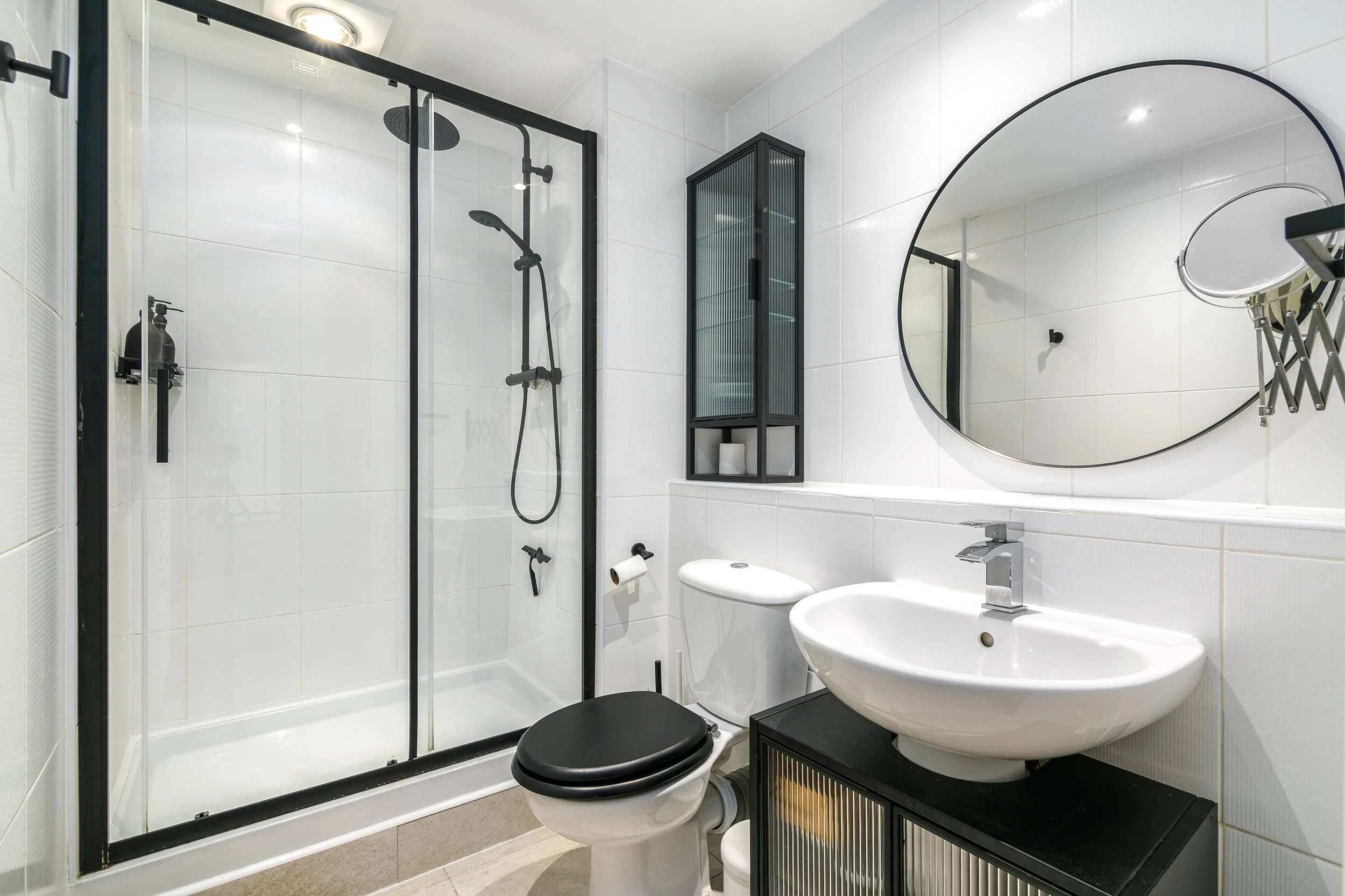 Modern bathroom with white tiled walls, a glass shower enclosure with black framing, a black showerhead and faucet, a black storage cabinet, a round mirror, a small sink, and a toilet with a black seat.