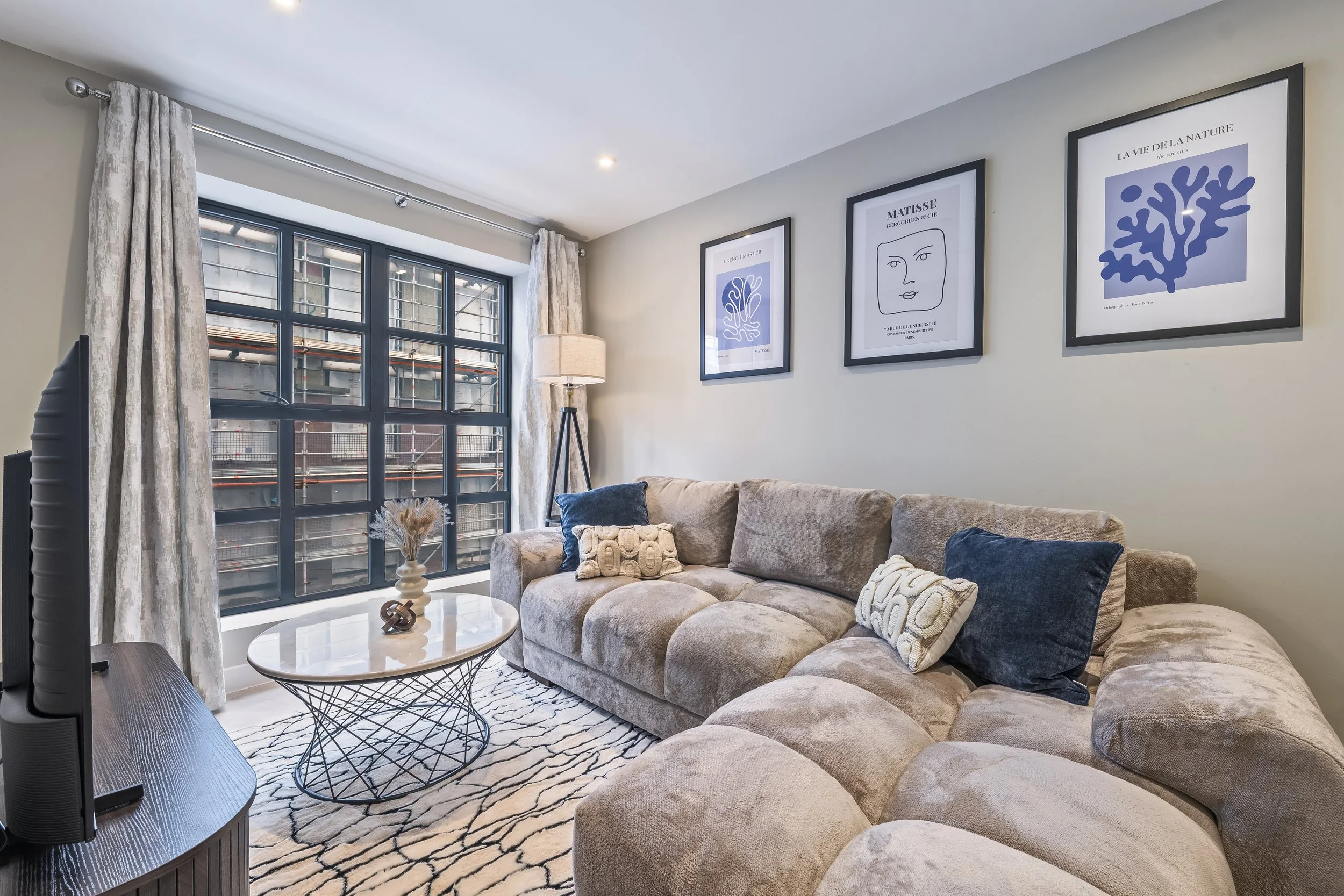 Living room with beige sectional sofa, decorative pillows, round white coffee table with black wire base, patterned rug, large window with metal burglar bars, framed modern artwork on light-colored wall, floor lamp, curtains, and a TV in the corner.