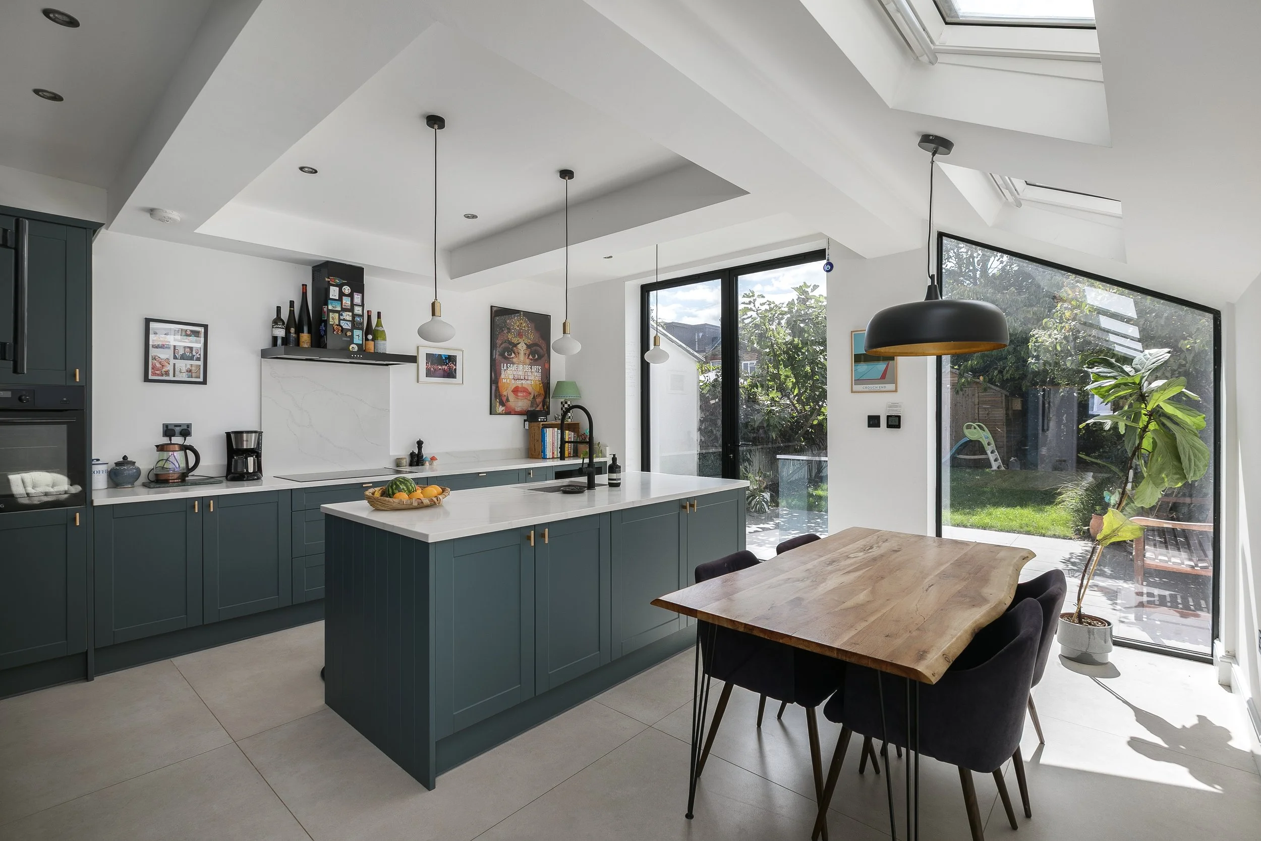 Modern kitchen with teal cabinets, white countertops, and a wooden dining table with black chairs. Large windows and skylights bring in natural light. Decor includes framed artwork, bottles, and plants.