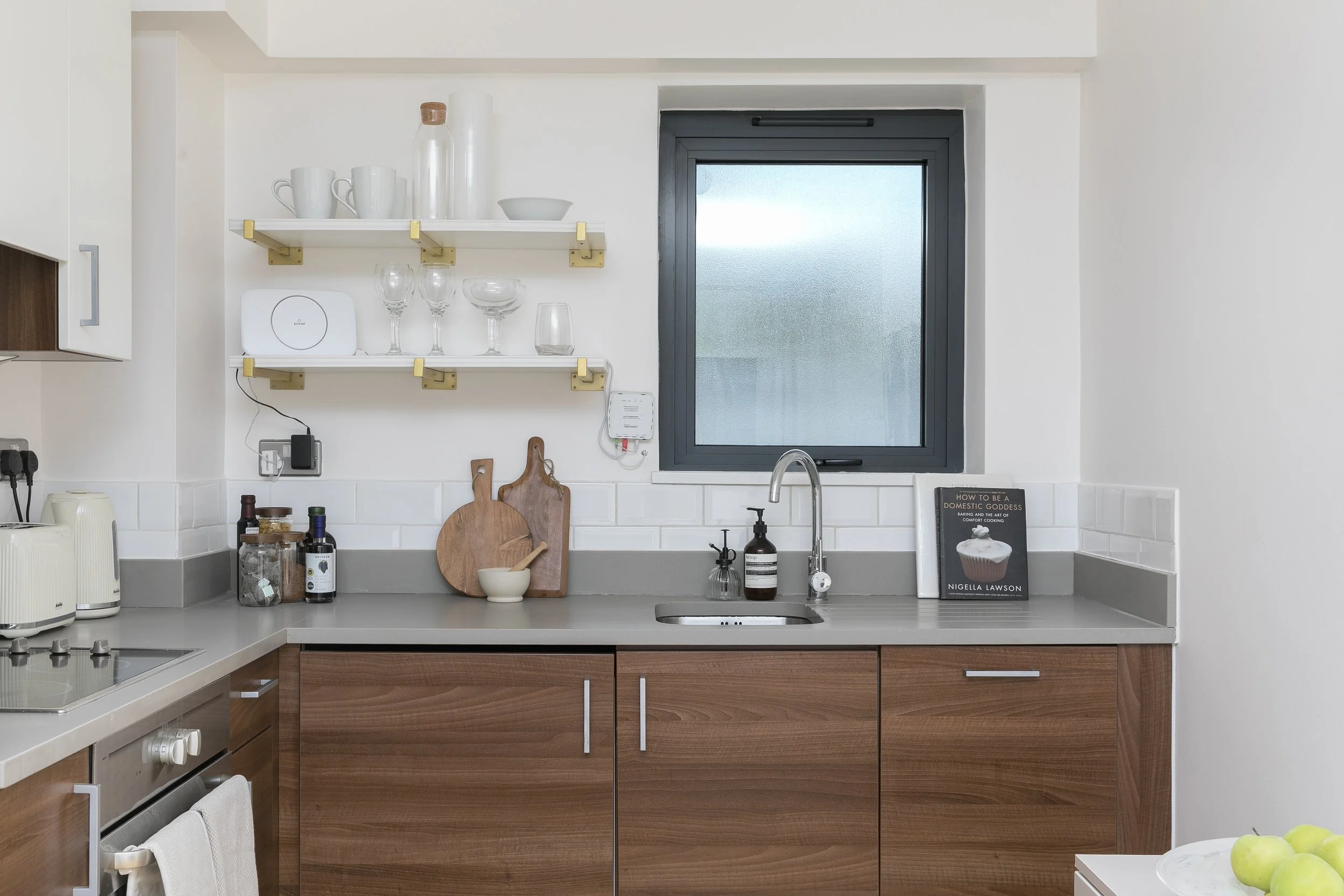 Modern kitchen with open shelving holding white mugs, glassware, and jars; window above sink; countertop with kitchen items and a cookbook.