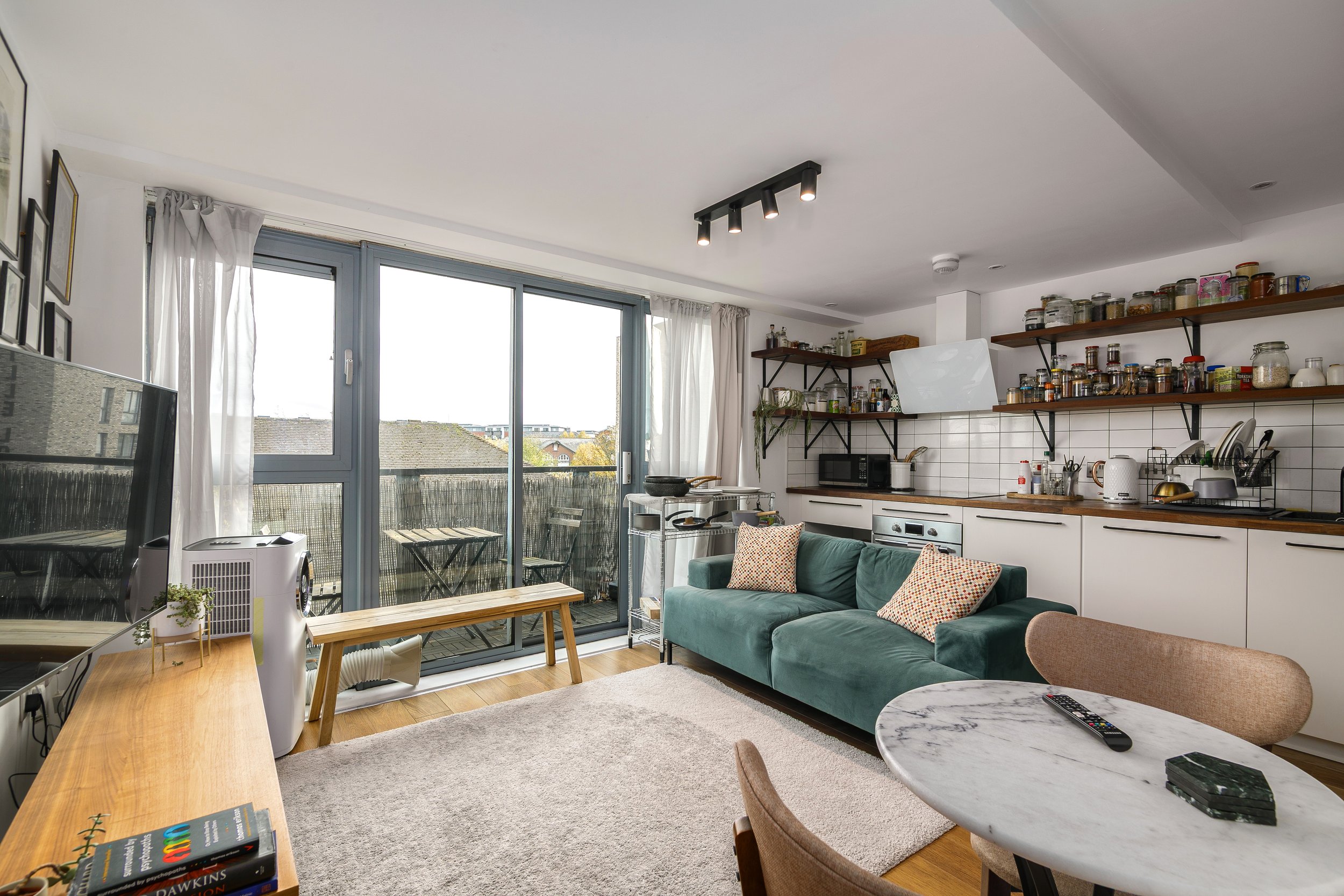 A modern apartment living room with large windows, a green sofa with patterned cushions, wooden and marble tables, open kitchen with shelves filled with jars, and a balcony with outdoor seating.