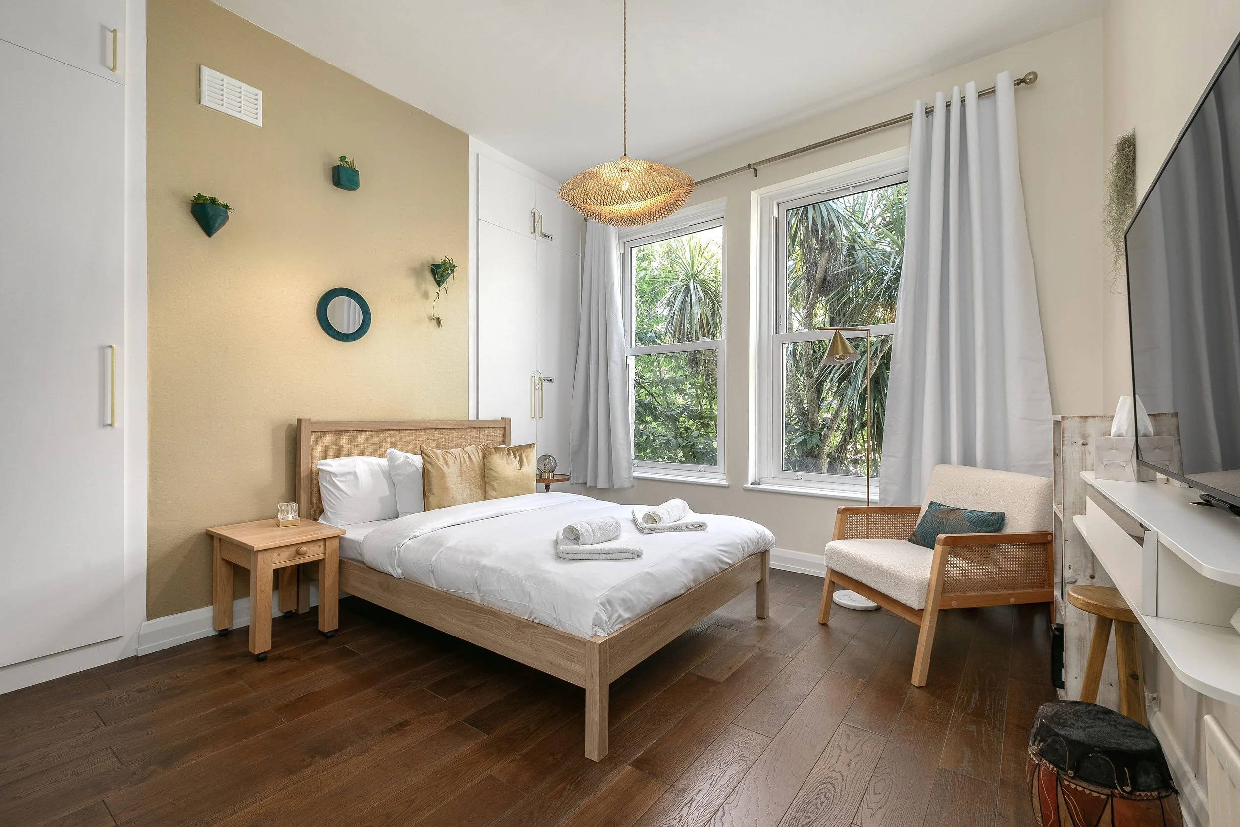 A cozy bedroom with cream-colored walls, a wooden bed with white bedding, and two folded towels on the bed. There are two large windows with white curtains, showing trees outside. Decor includes a round mirror, small wall planters, and a woven ceilin
