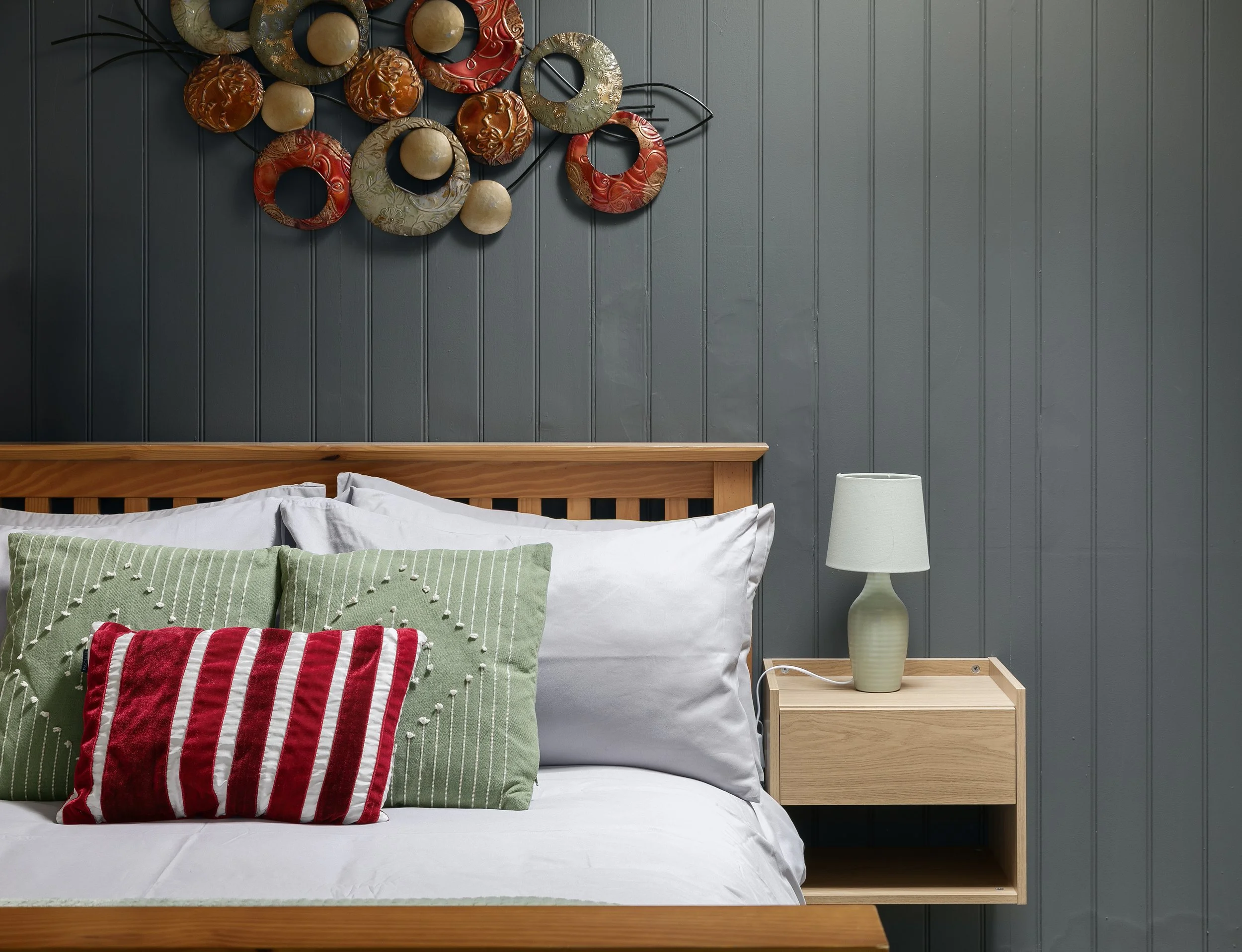 A bedroom with a wooden bed frame, decorated with green and red pillows, a gray bedside table with a lamp, and a gray paneled wall with abstract circular metal art.