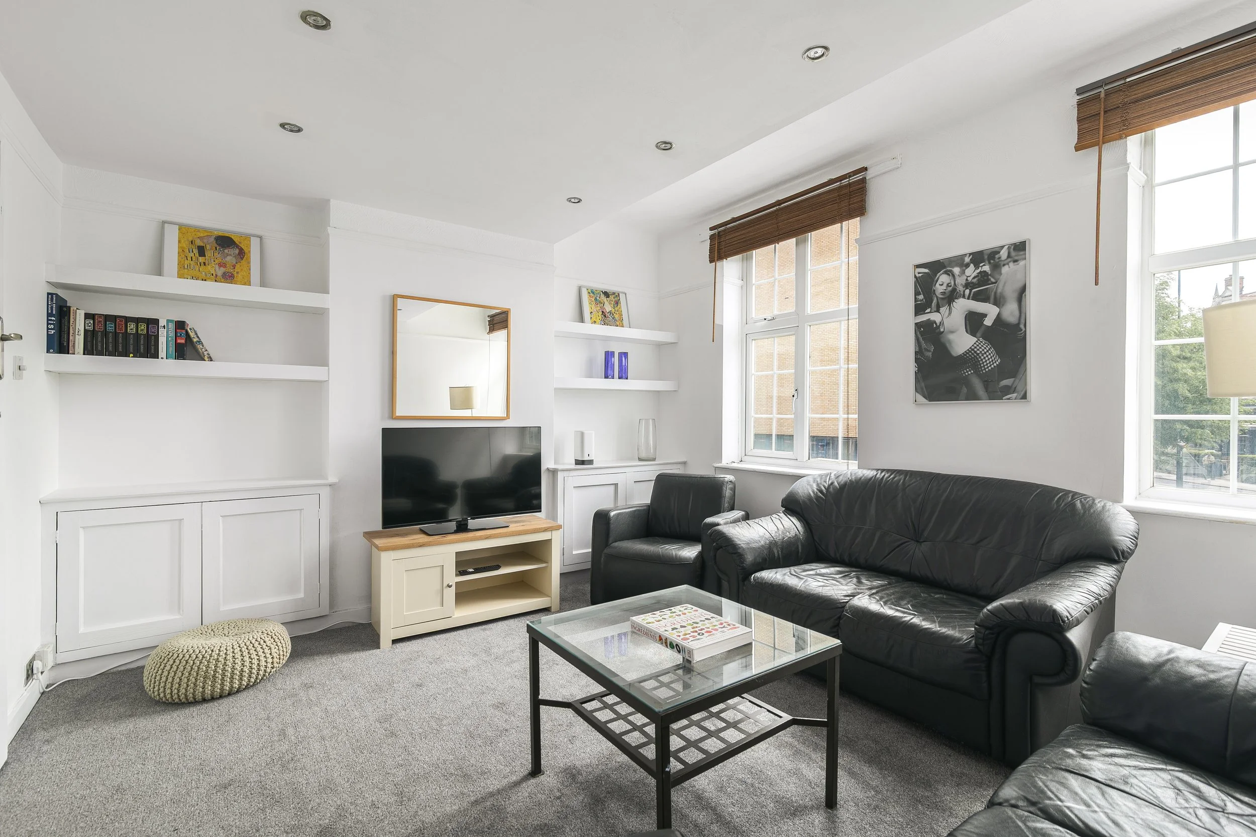 A living room with white walls, large windows, black leather couch, coffee table, bookshelves, and wall art.