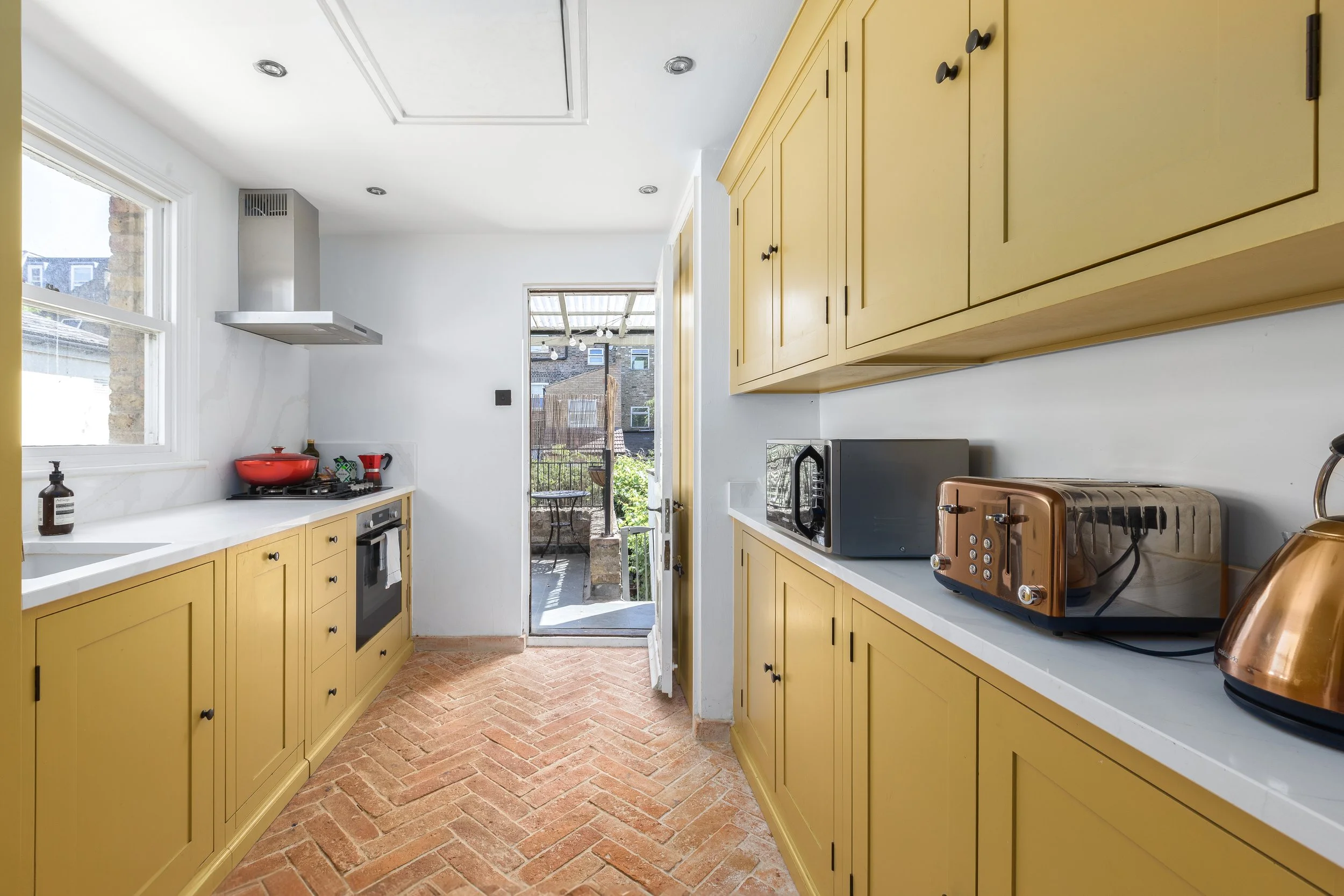 A kitchen with yellow cabinets, white countertops, a brick floor, and a door leading to an outdoor patio. There is a red pot on the stove, a microwave, a toaster, and a copper kettle on the counter.