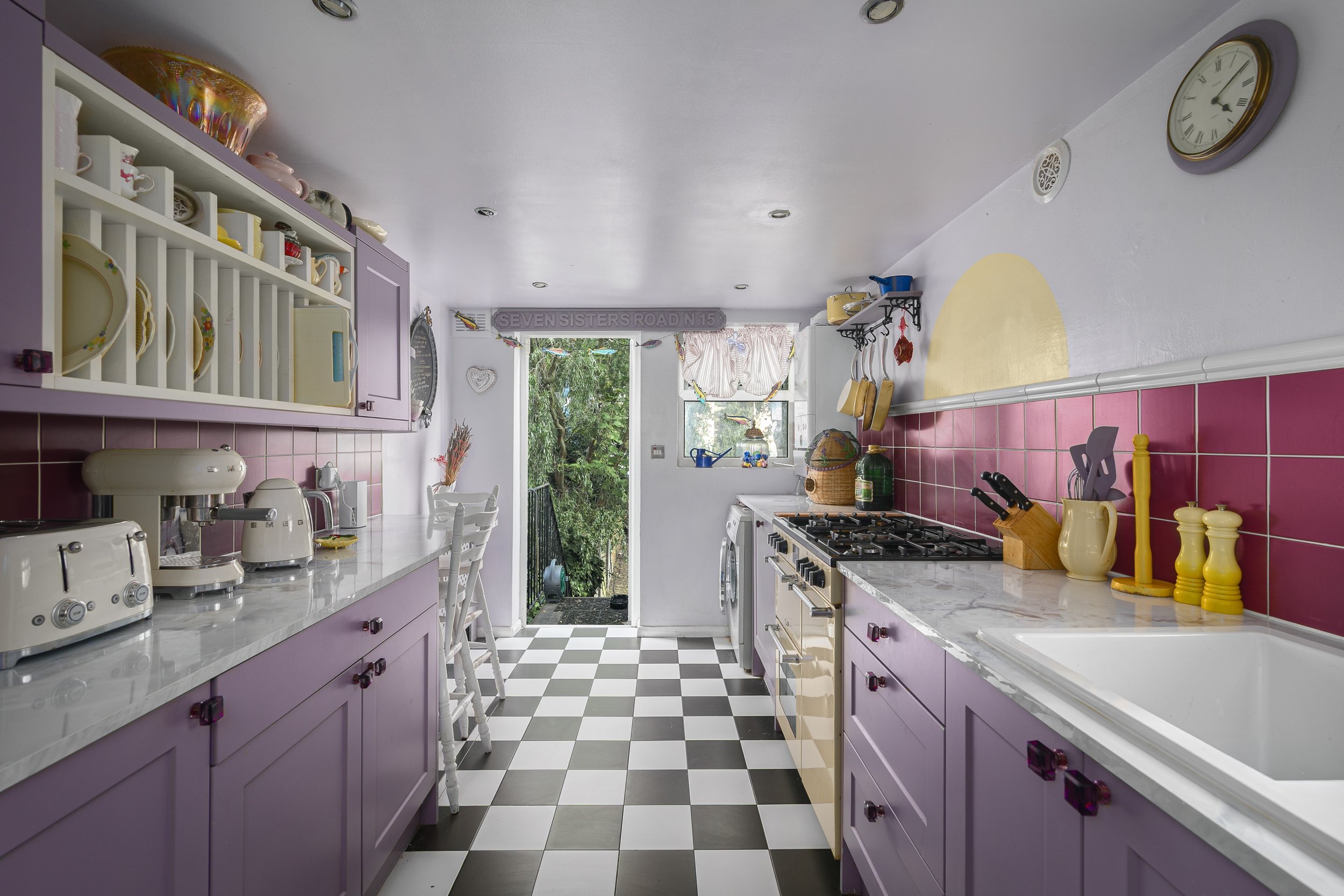 A colorful kitchen with purple cabinets, white marble countertops, a black and white checkered floor, and pink tiled backsplash. There are various kitchen appliances, utensils, and decor, with a window showing greenery outside.