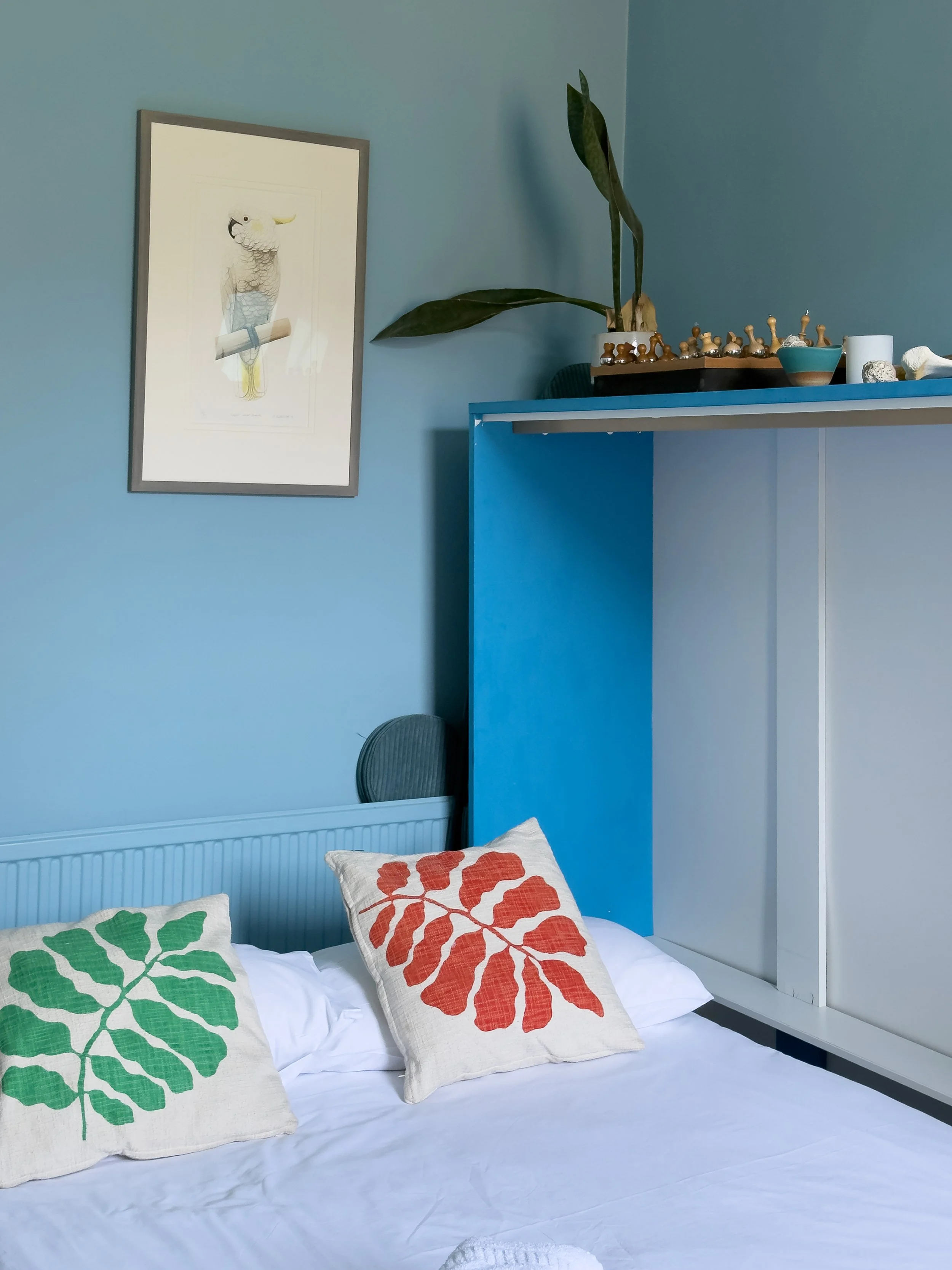 A neatly made bed with white linens and three decorative pillows featuring orange, green, and red leaf patterns. The bed is against a blue wall with a framed illustration of a bird perched on a branch to the left, and a wall with a built-in shelf holding a tall plant with large green leaves, chess pieces, and small decorative items.