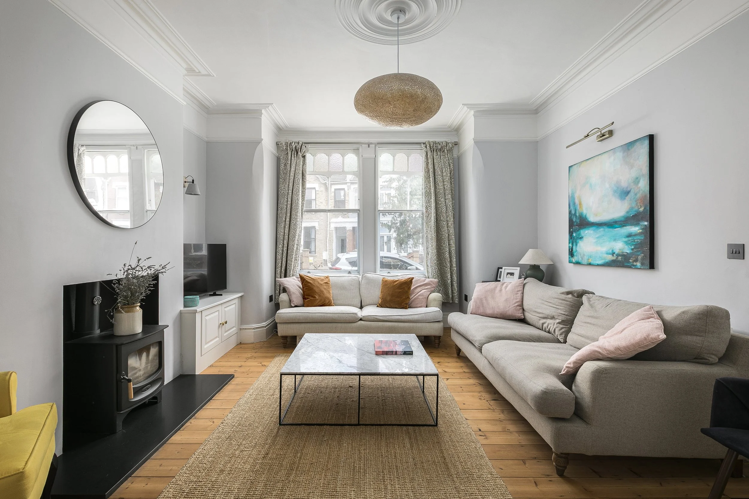 Living room with white walls, a large window with patterned curtains, two sofas with pillows, a coffee table, a wall-mounted painting, a mirror, and a fireplace, with a ceiling light fixture.