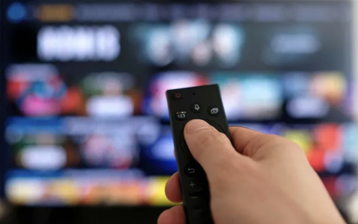 Close-up of a hand holding a TV remote pointing at a blurred television screen displaying a streaming service menu.