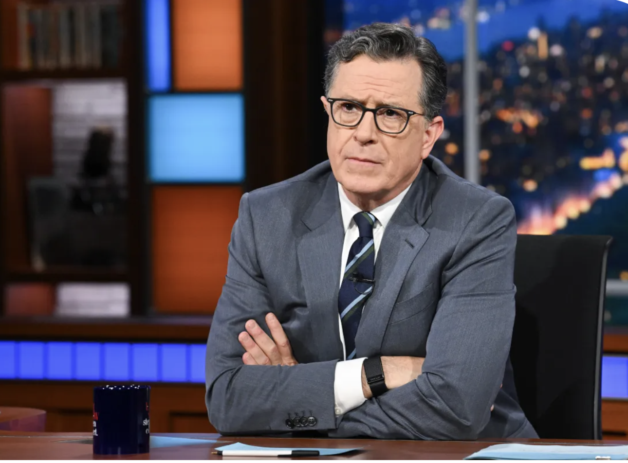 A man in a gray suit and glasses sitting at a TV studio desk with crossed arms, facing the camera with a serious expression.