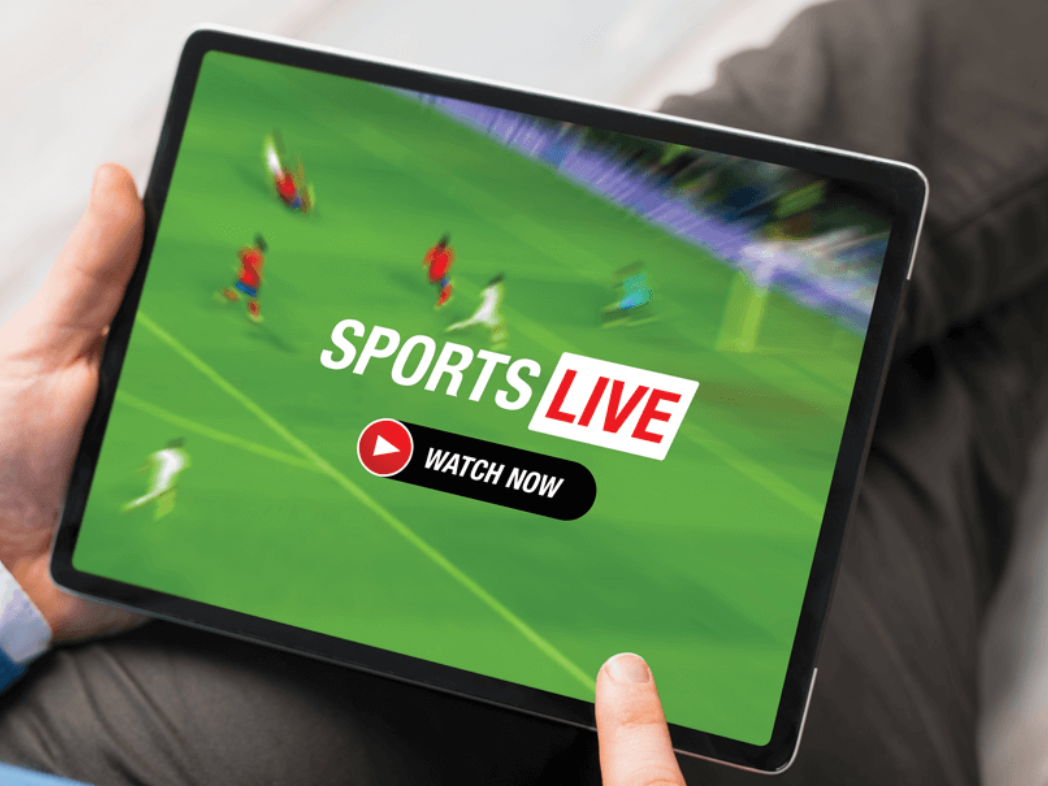 Person holding a tablet displaying a live sports broadcast with a green field and players, with an overlay that says "SPORTS LIVE" and a "WATCH NOW" button.