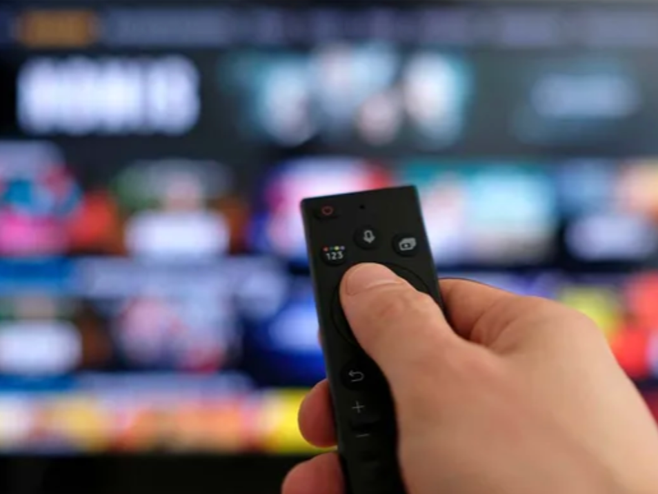 Close-up of a hand holding a TV remote control in front of a blurred television screen with multiple app icons.
