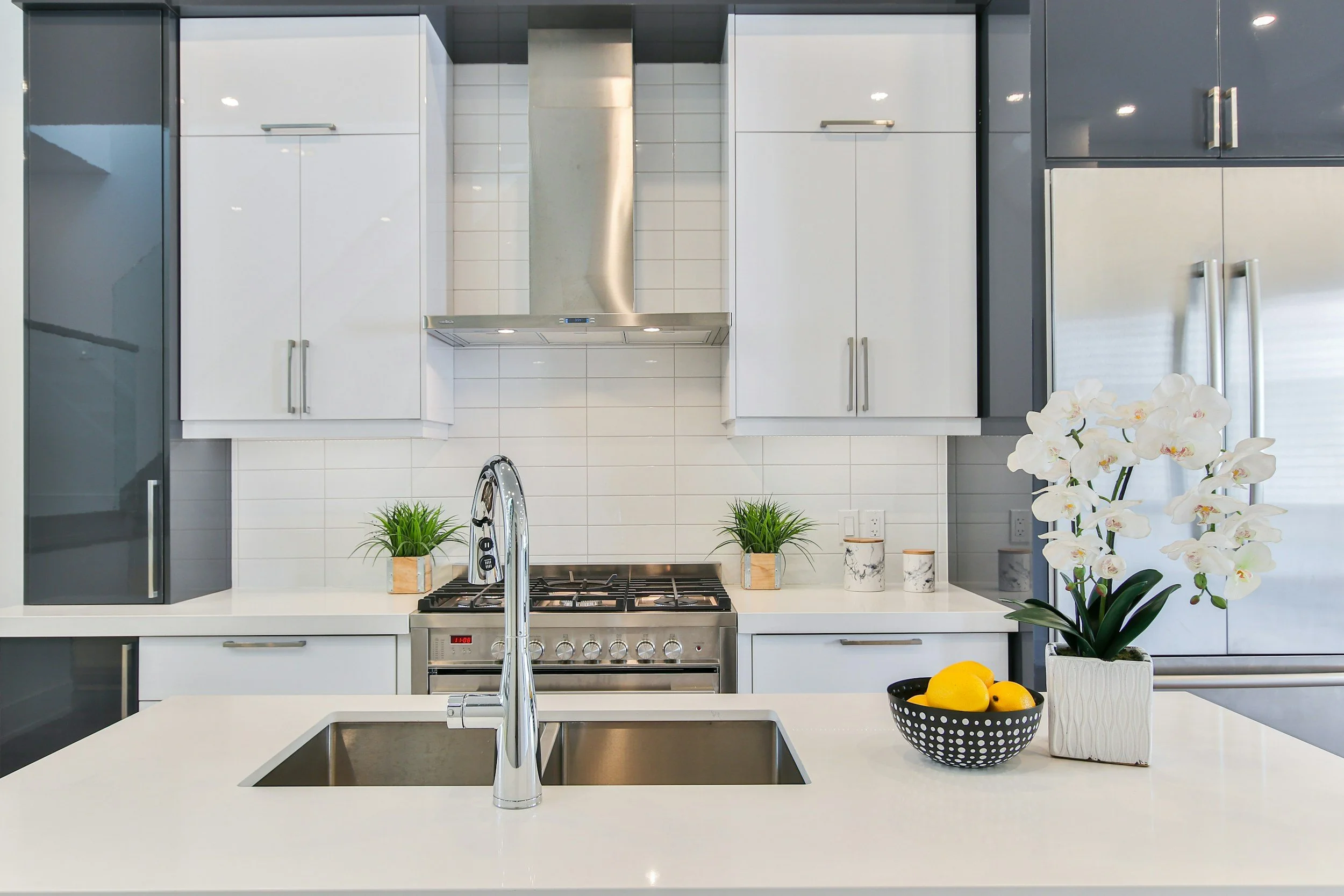 Modern kitchen with white countertops, stainless steel appliances, white tile backsplash, dark gray and white cabinetry, decorated with potted plants, white orchids, and a black bowl of lemons.