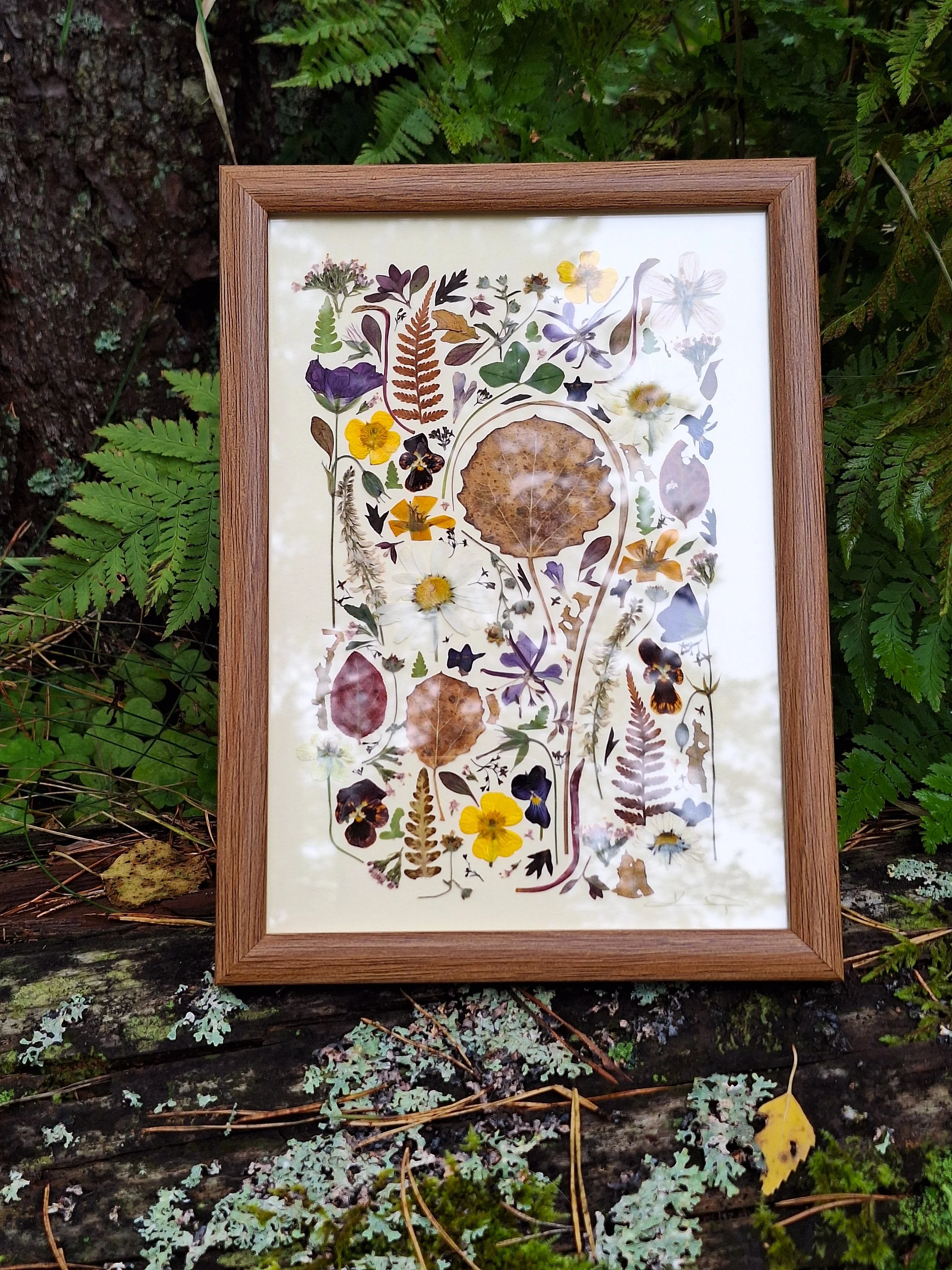 A framed pressed flower mosaic artwork by Kai Lillekaardid. The piece features a dense arrangement of colorful wildflowers, ferns, and a large central brown aspen leaf in a warm wood frame.
