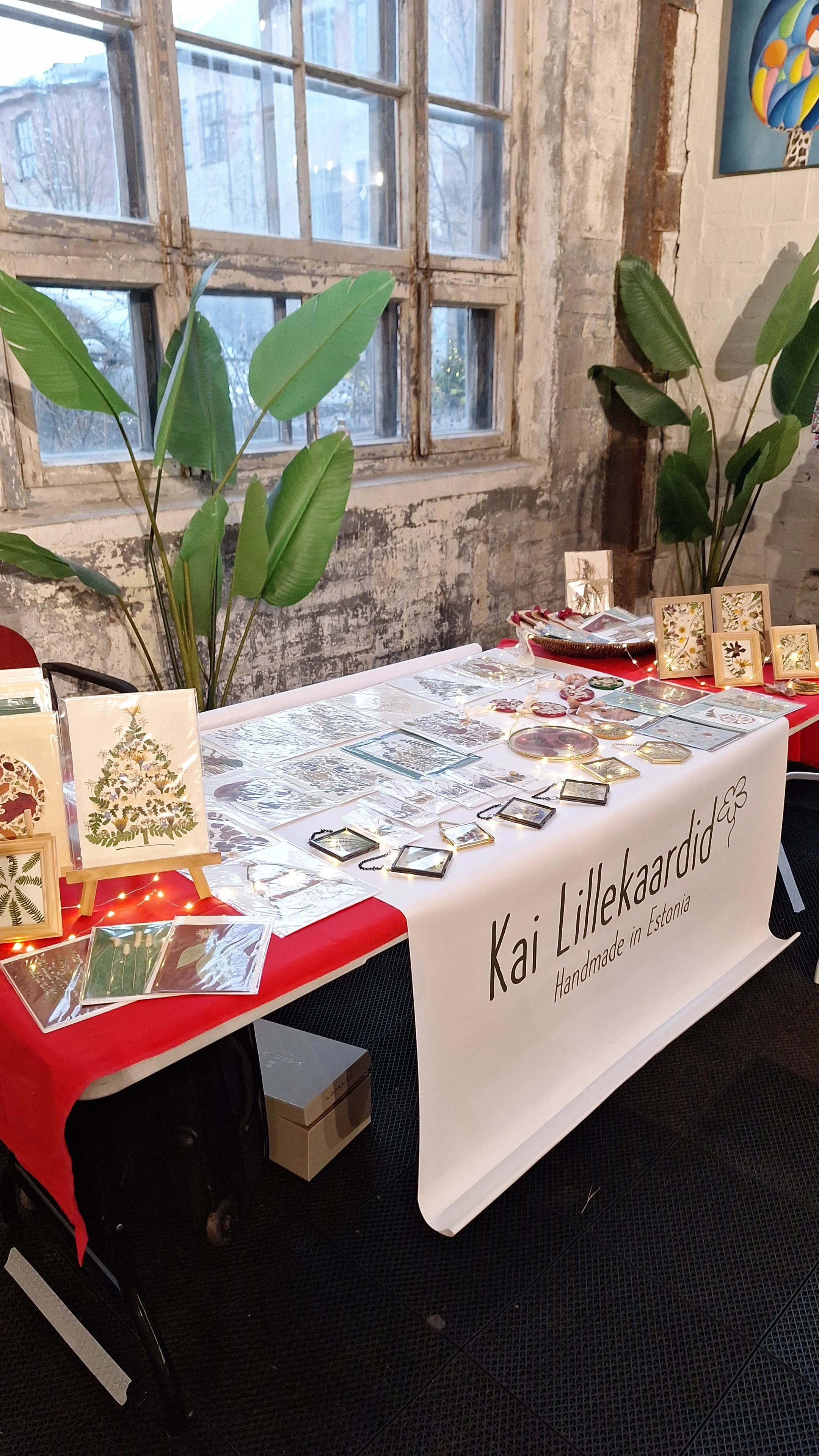 A table display at an indoor market with various framed artwork and jewelry, decorated with string lights, against a rustic brick wall with large green plants nearby.