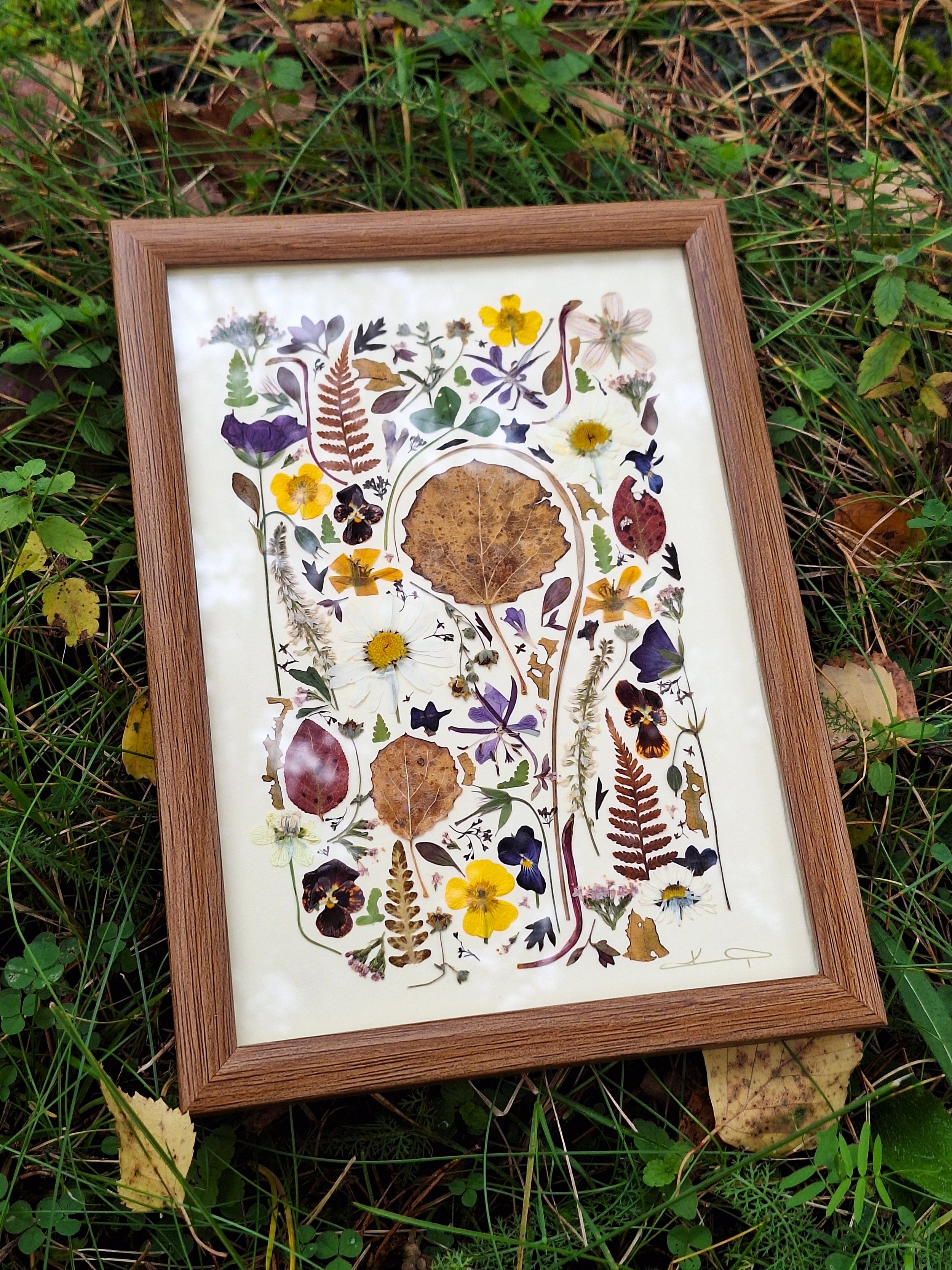 A framed pressed flower mosaic artwork by Kai Lillekaardid. The piece features a dense arrangement of colorful wildflowers, ferns, and a large central brown aspen leaf in a warm wood frame.