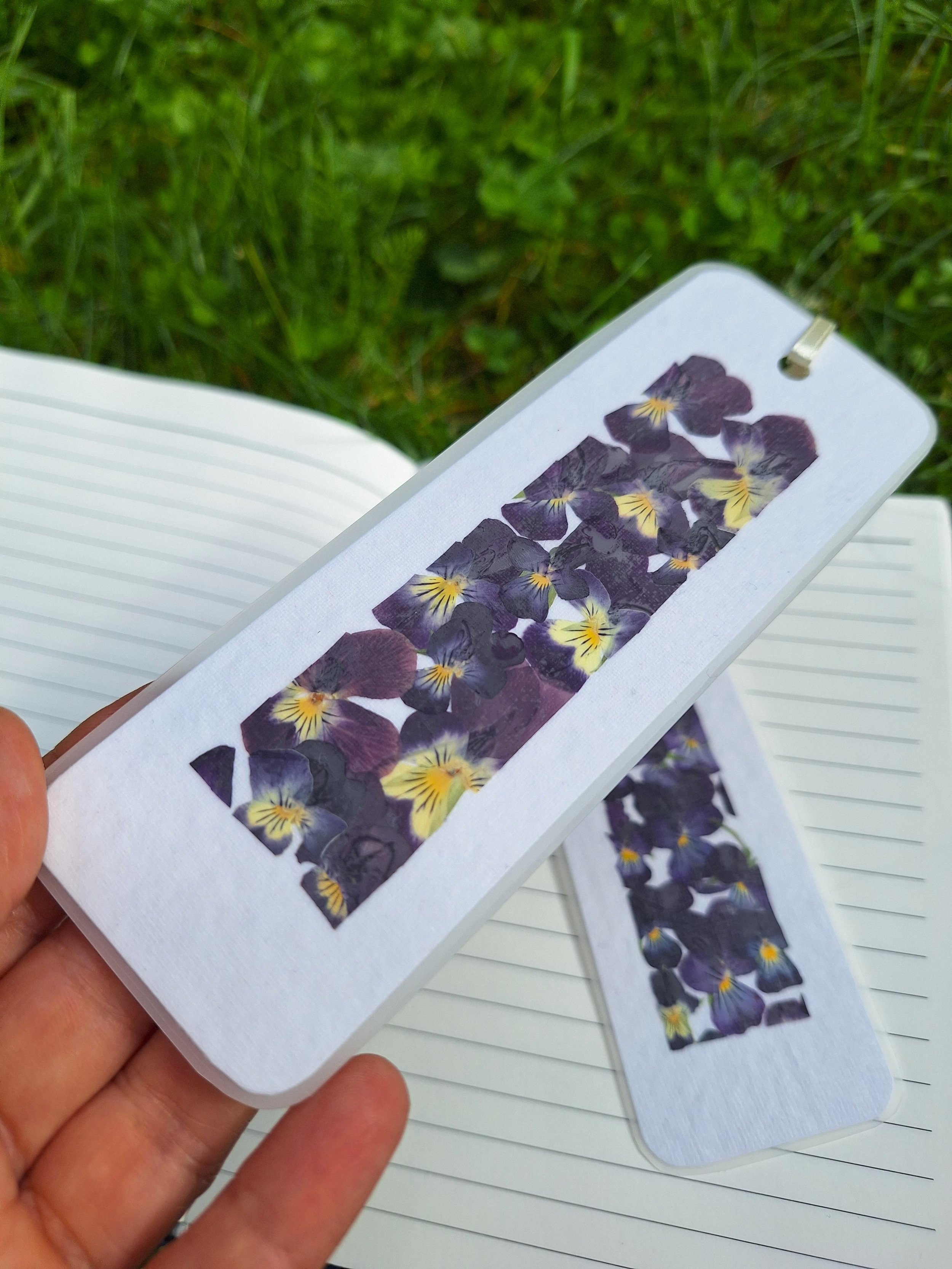 Two laminated bookmarks side-by-side featuring bright yellow and deep velvet purple pressed flowers carefully laid out on handmade textured paper.