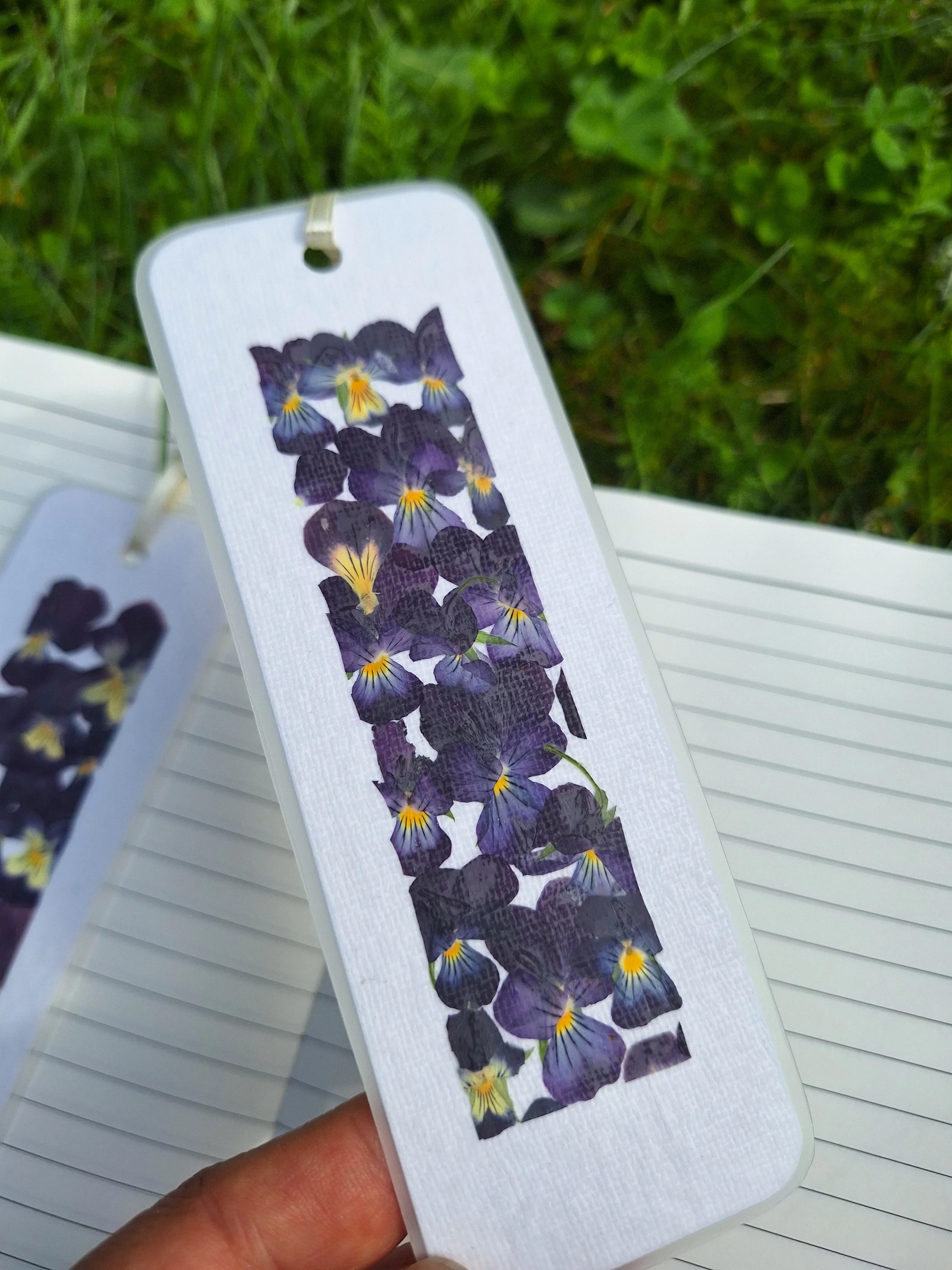 Two laminated bookmarks side-by-side featuring bright yellow and deep velvet purple pressed flowers carefully laid out on handmade textured paper.