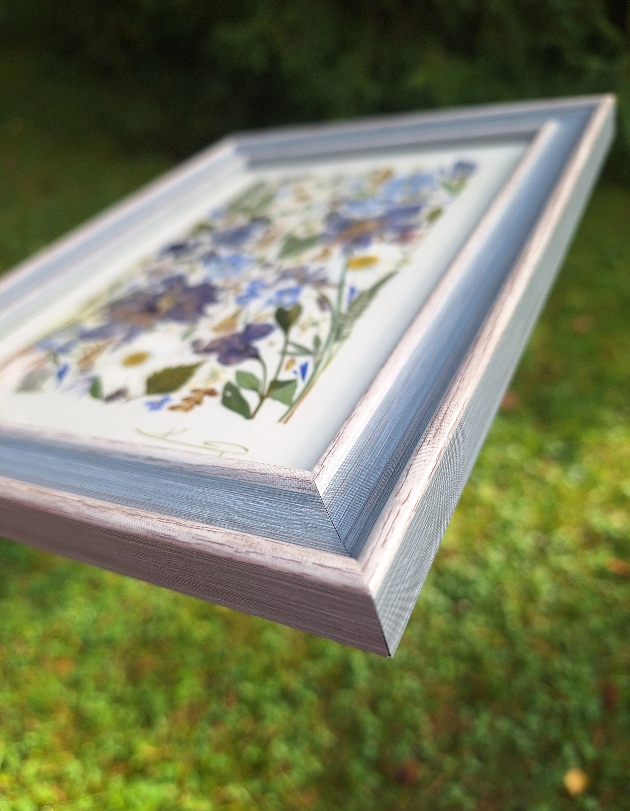 Angled view of an authentic pressed wildflower art frame by Kai Lillekaardid featuring a vertical arrangement of blue columbines, white daisies, and purple pansies in a light grey-blue wooden frame.
