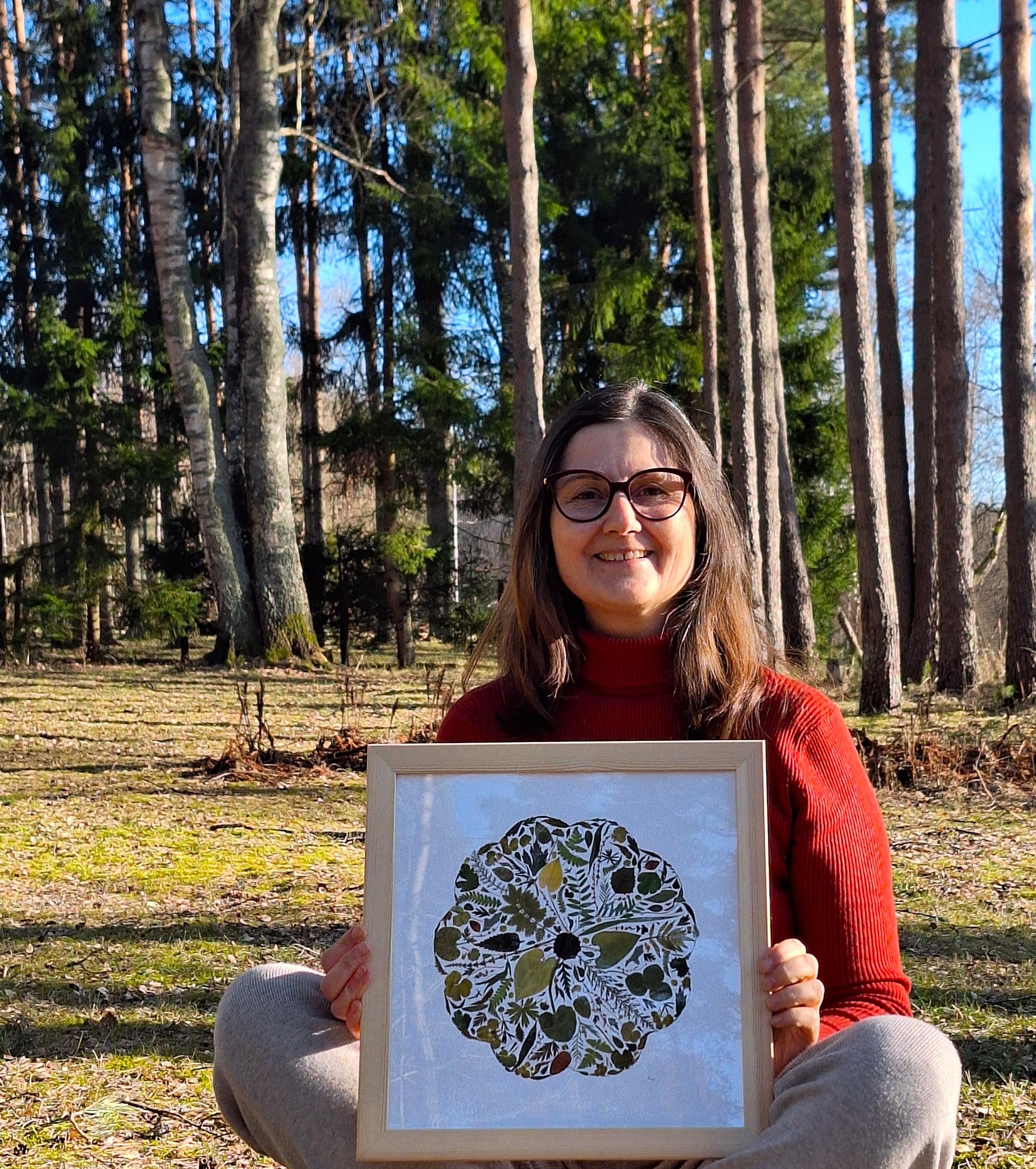 A woman sitting outdoors in a wooded area holds a framed picture of a skull art print made of leaves and plant patterns. She is wearing glasses and a red sweater, smiling at the camera.