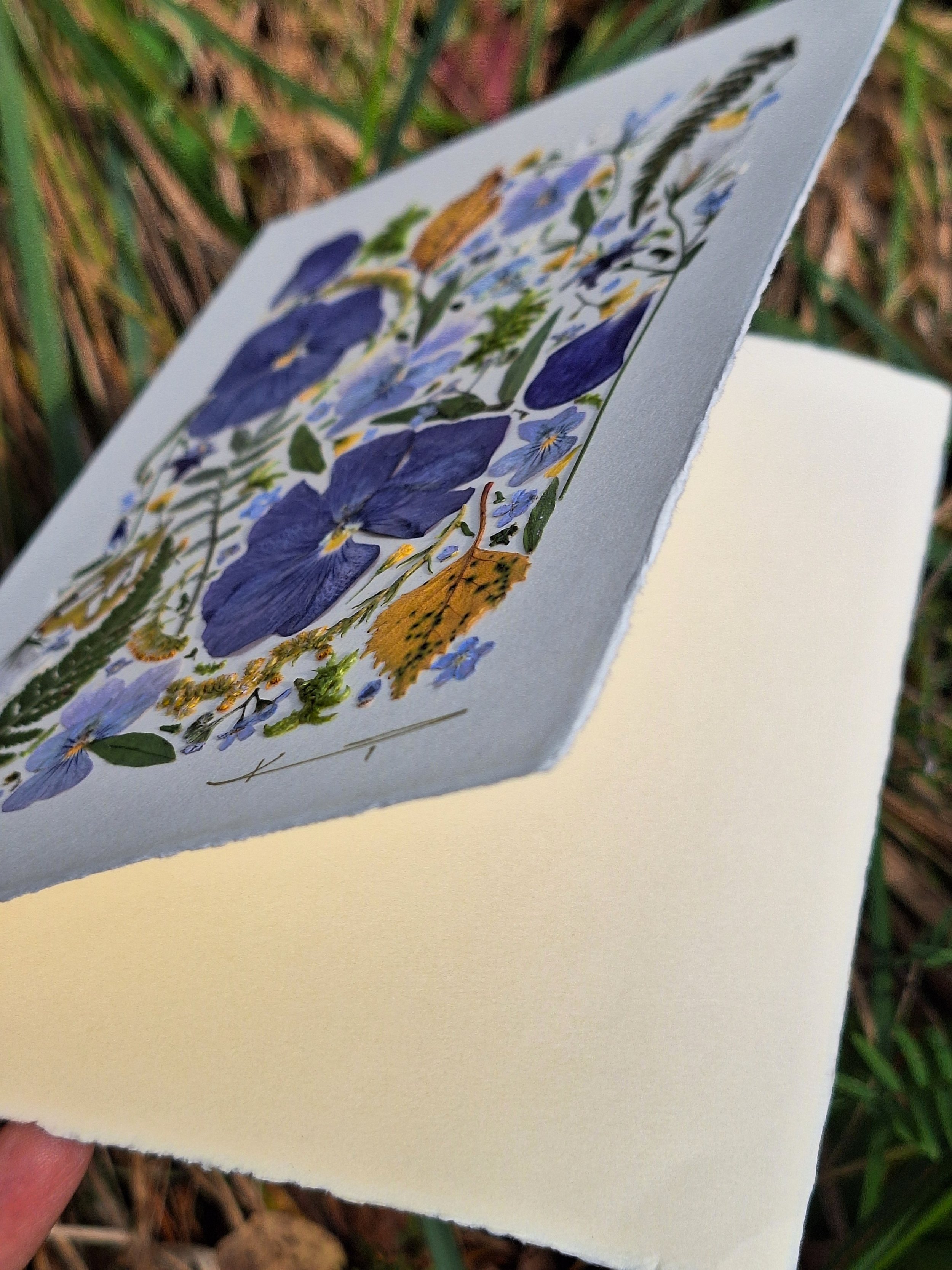 Kai Lillekaardid handmade seasonal greeting card titled The Indigo Equinox, featuring indigo pressed pansies, yellow autumn leaves, and green ferns displayed on a small wooden easel in nature.