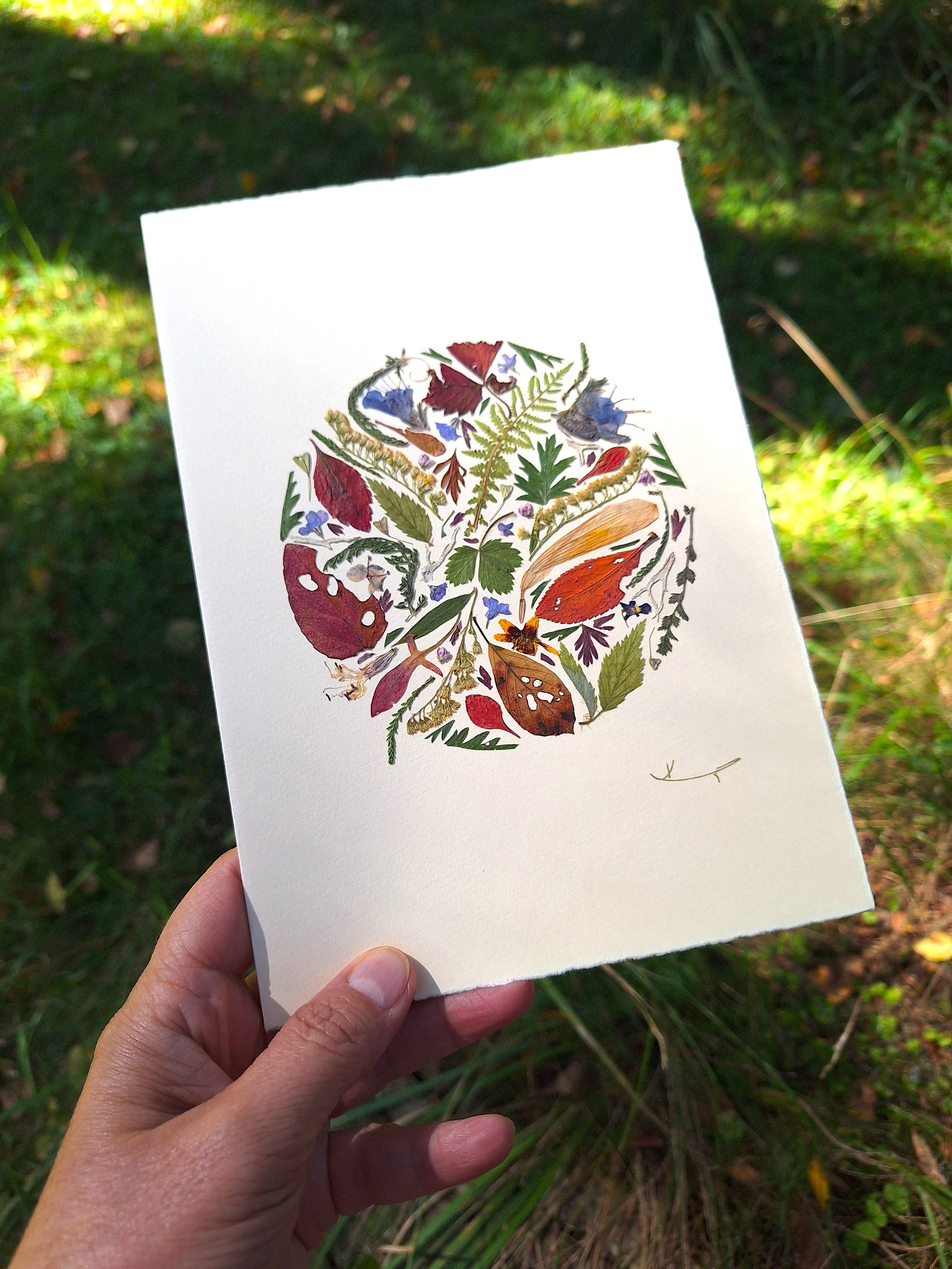 Side angle view of an authentic pressed leaf greeting card by Kai Lillekaardid showing an intricate circular arrangement of autumn ferns, green leaves, and brown foliage on handmade paper.