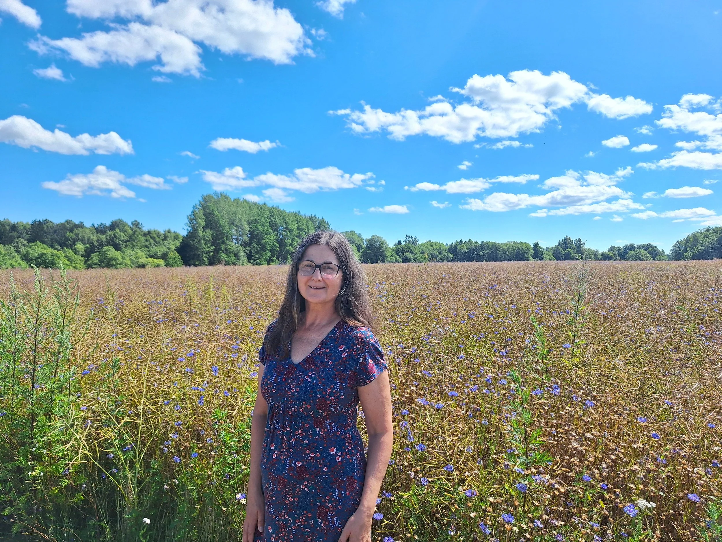 A woman with glasses and long hair standing in a purple flower field under a blue sky with scattered clouds.