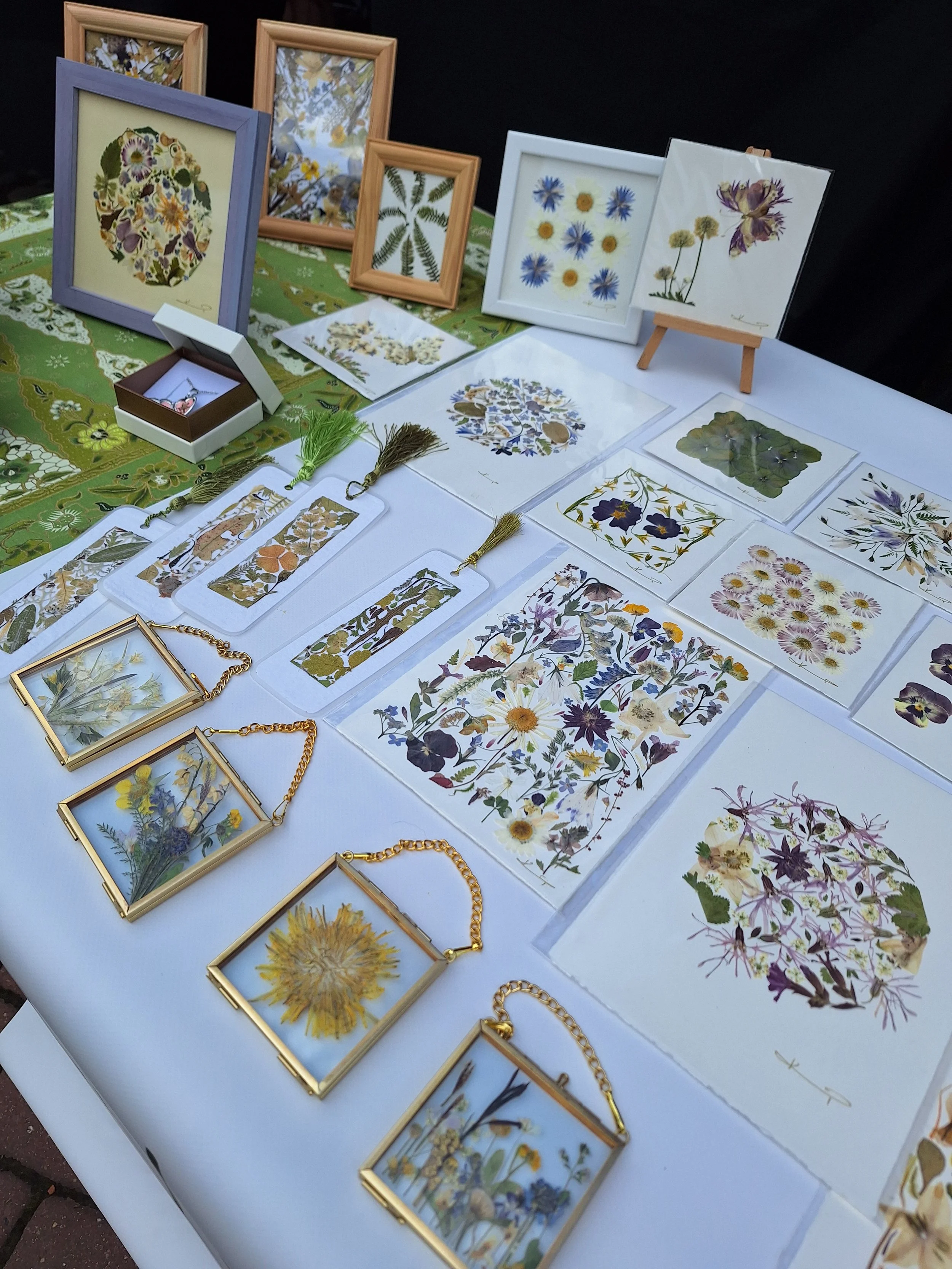 Display of pressed and framed botanical illustrations, jewelry, and art pieces on a table at an outdoor market.