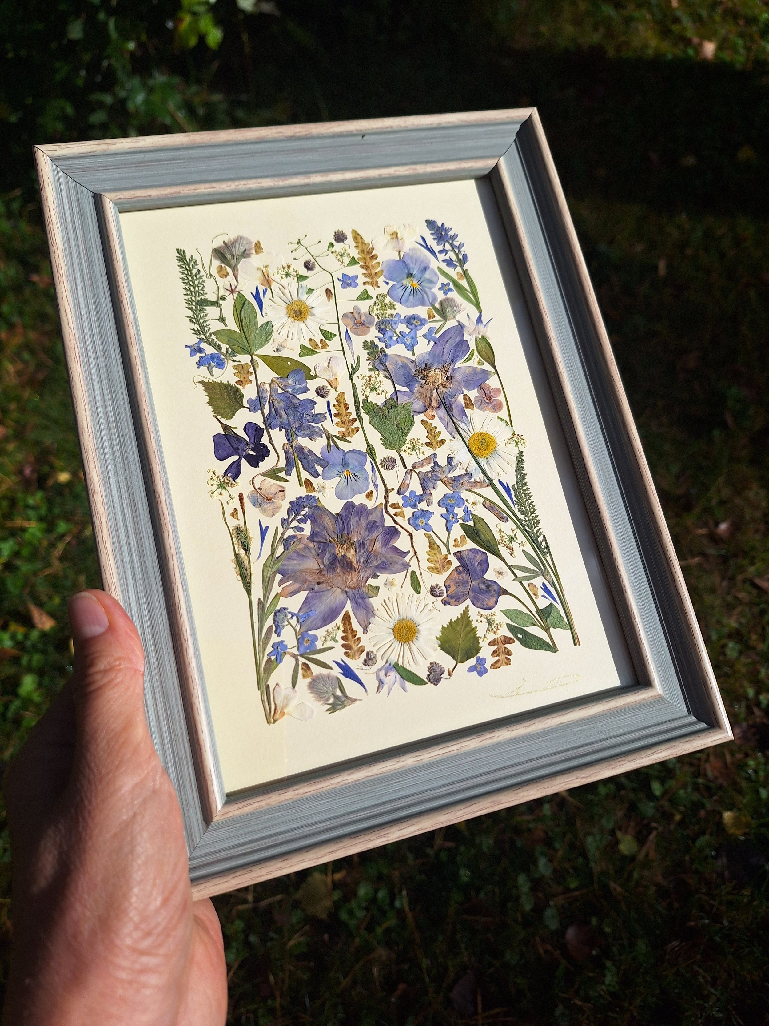 Angled view of an authentic pressed wildflower art frame by Kai Lillekaardid featuring a vertical arrangement of blue columbines, white daisies, and purple pansies in a light grey-blue wooden frame.