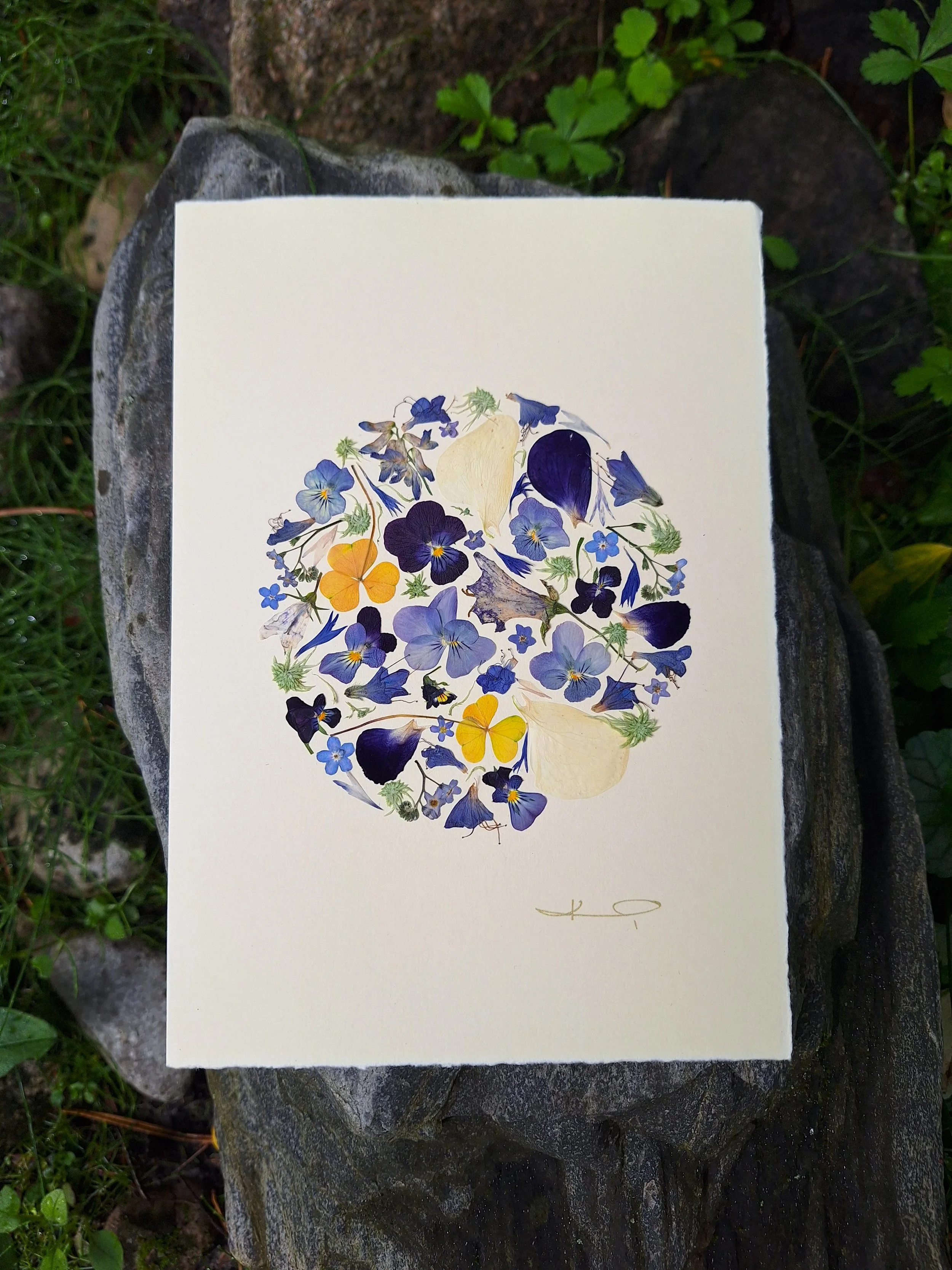 Close-up of a handmade greeting card by Kai Lillekaardid showcasing a circular pressed flower mosaic with light blue flowers, midnight violet petals, and yellow accents on premium cream paper.