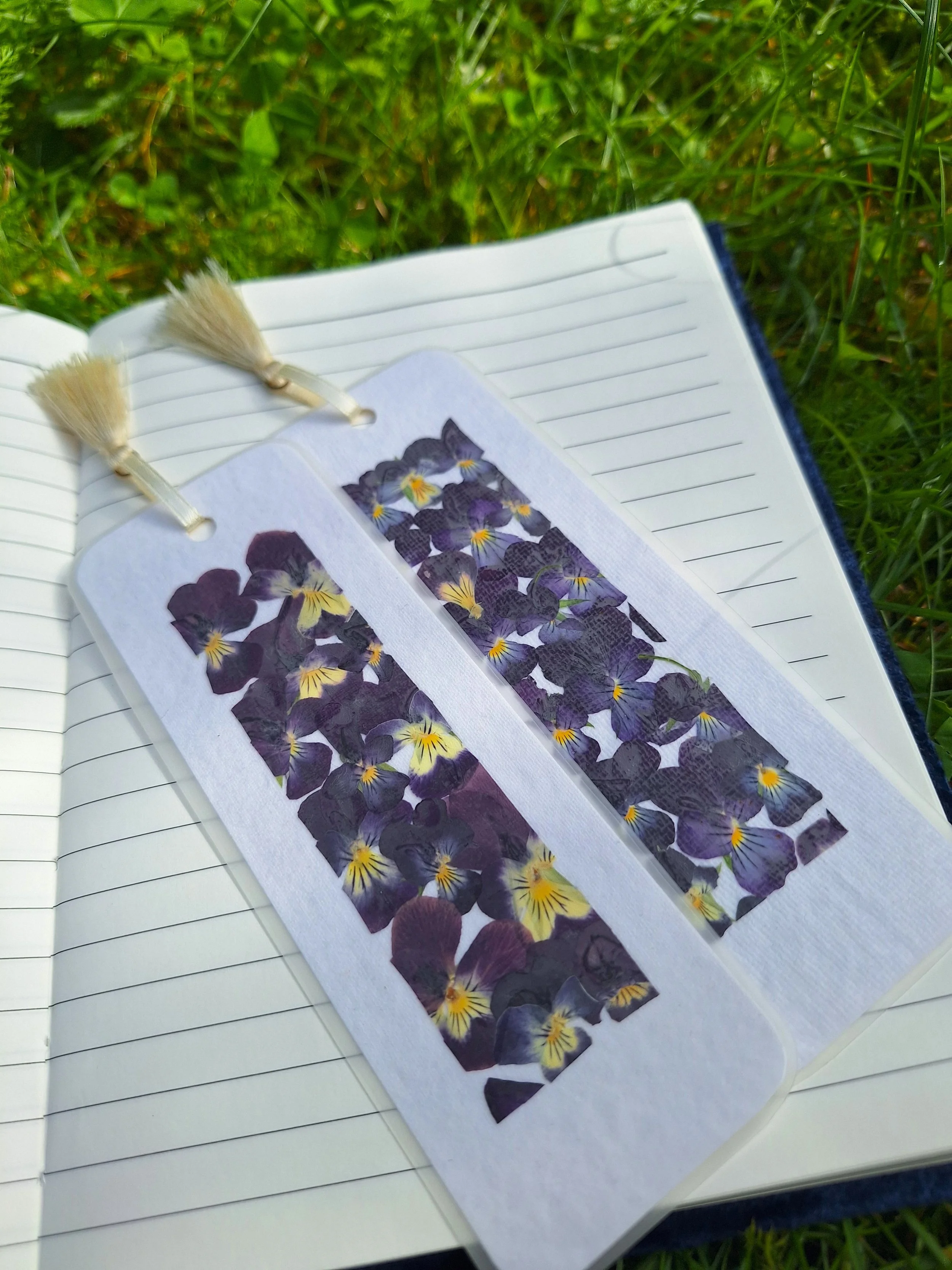 Two laminated bookmarks side-by-side featuring bright yellow and deep velvet purple pressed flowers carefully laid out on handmade textured paper.