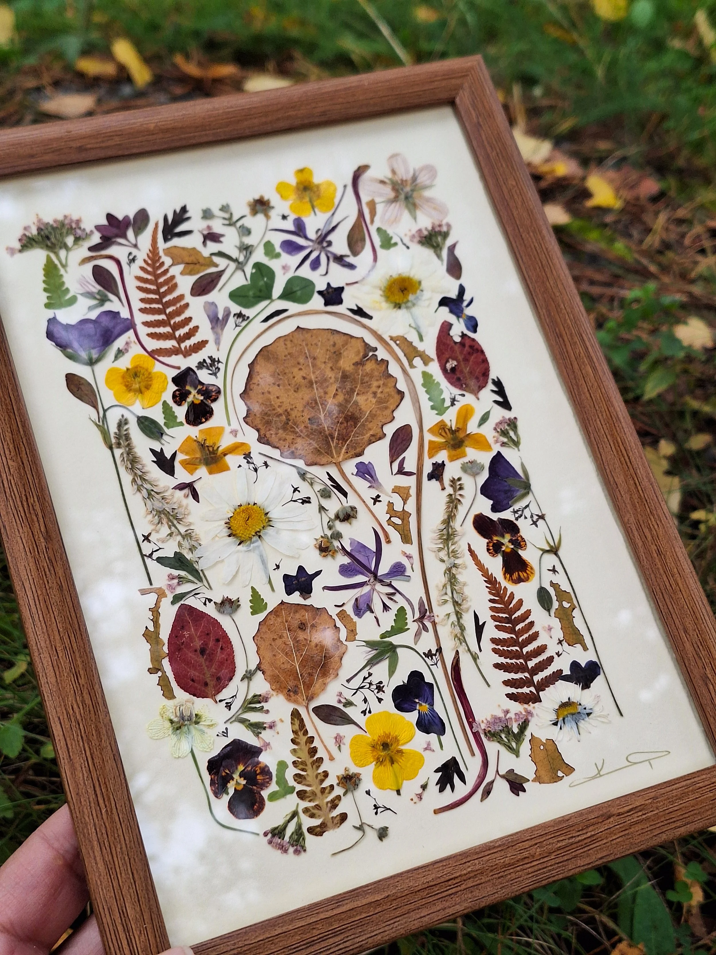 A framed pressed flower mosaic artwork by Kai Lillekaardid. The piece features a dense arrangement of colorful wildflowers, ferns, and a large central brown aspen leaf in a warm wood frame.