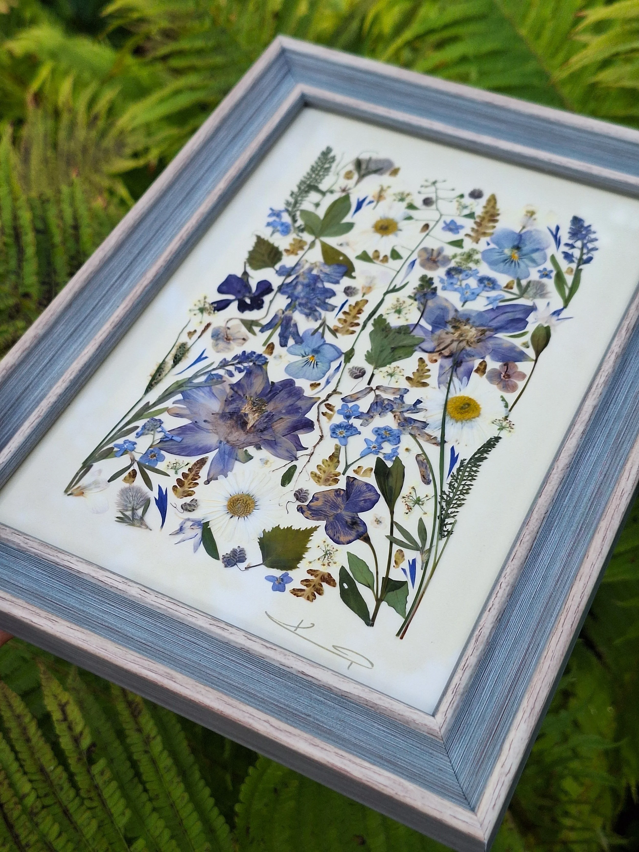 Angled view of an authentic pressed wildflower art frame by Kai Lillekaardid featuring a vertical arrangement of blue columbines, white daisies, and purple pansies in a light grey-blue wooden frame.