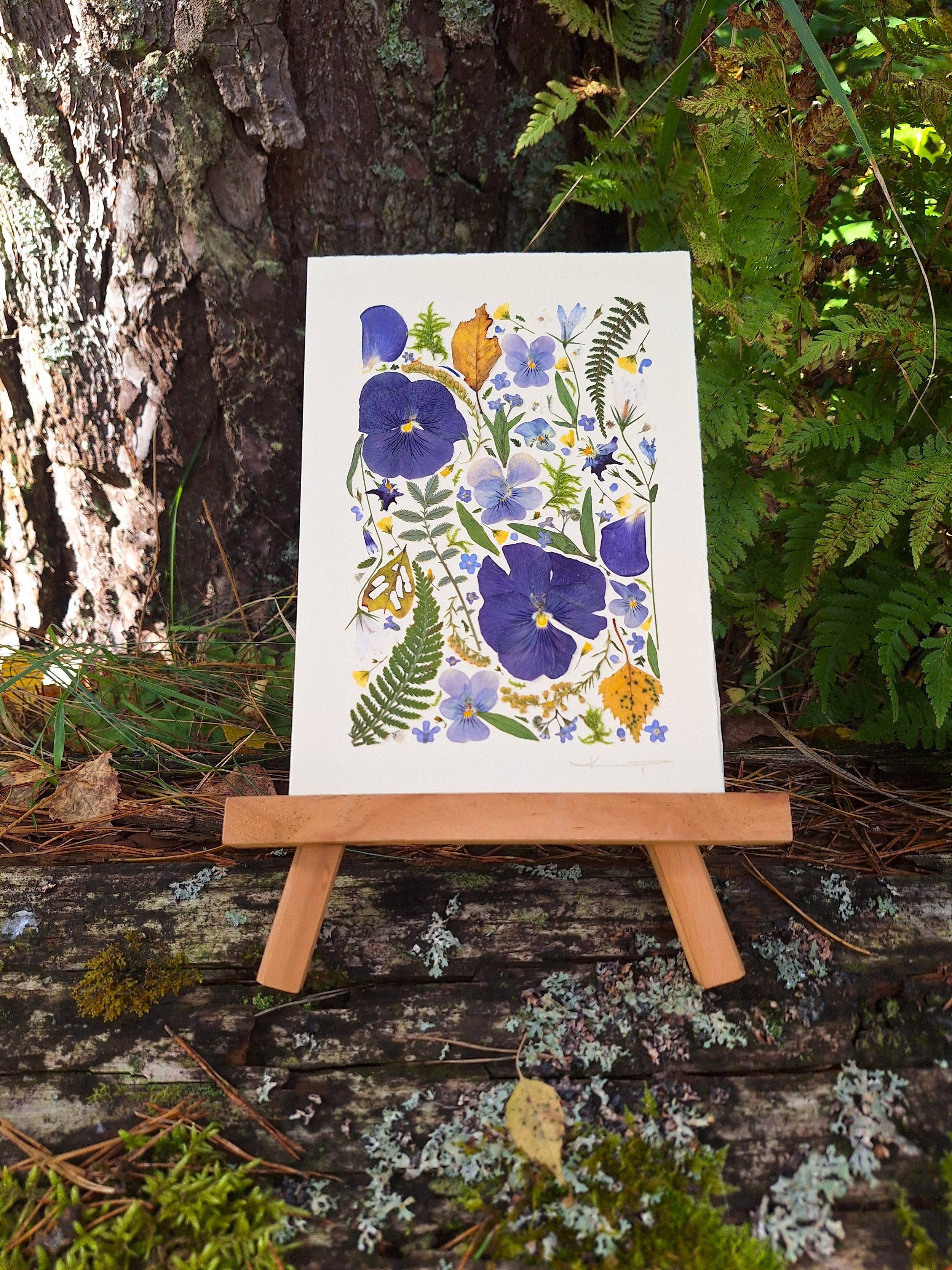 Kai Lillekaardid handmade seasonal greeting card titled The Indigo Equinox, featuring indigo pressed pansies, yellow autumn leaves, and green ferns displayed on a small wooden easel in nature.