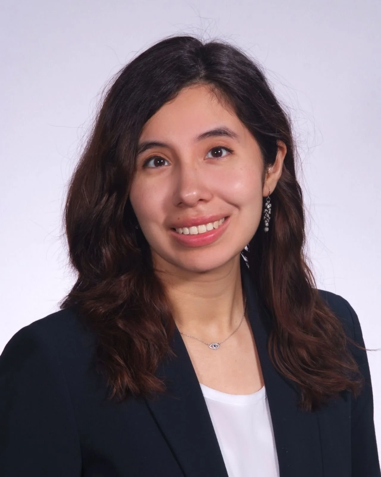 Headshot of a woman with long, wavy dark brown hair, wearing earrings, a necklace, a black blazer, and a white top, against a plain white background.