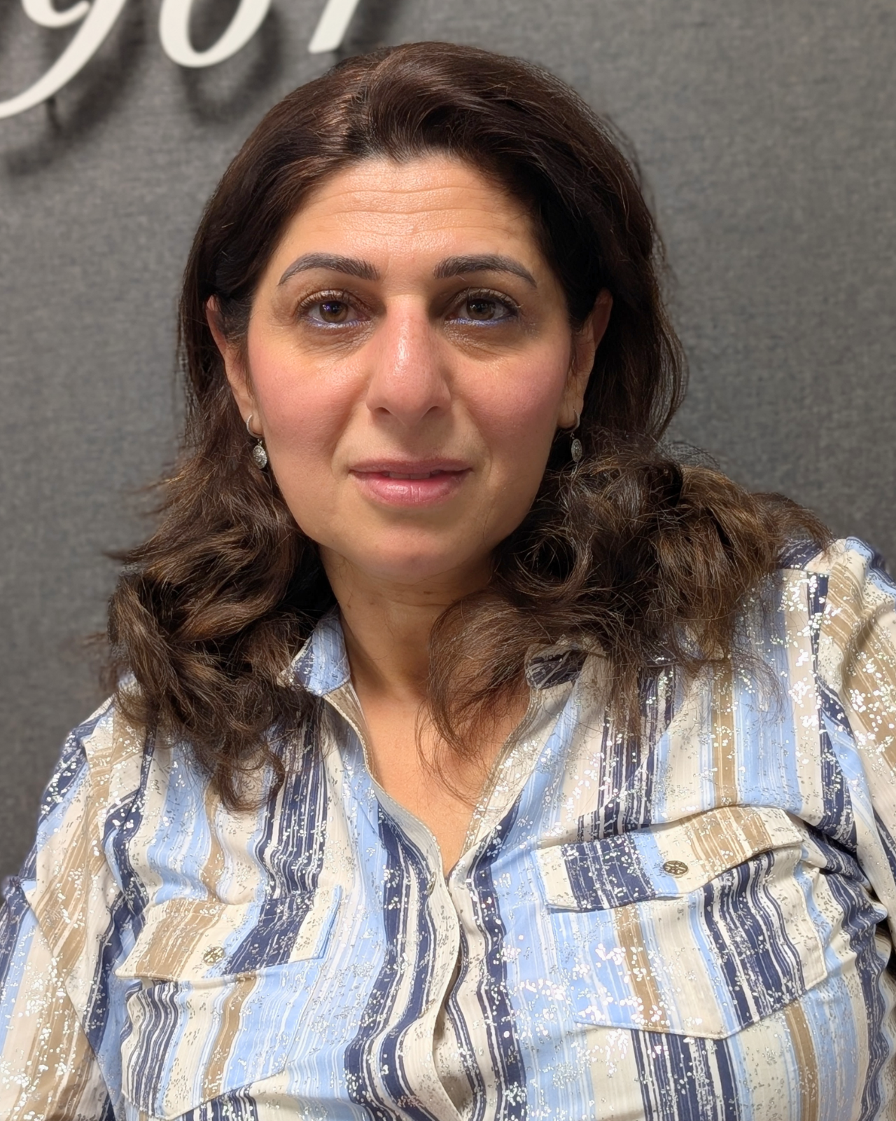 A woman with medium-length, wavy brown hair, wearing a patterned button-up shirt with blue, beige, and white colors, and earrings, standing in front of a gray background.