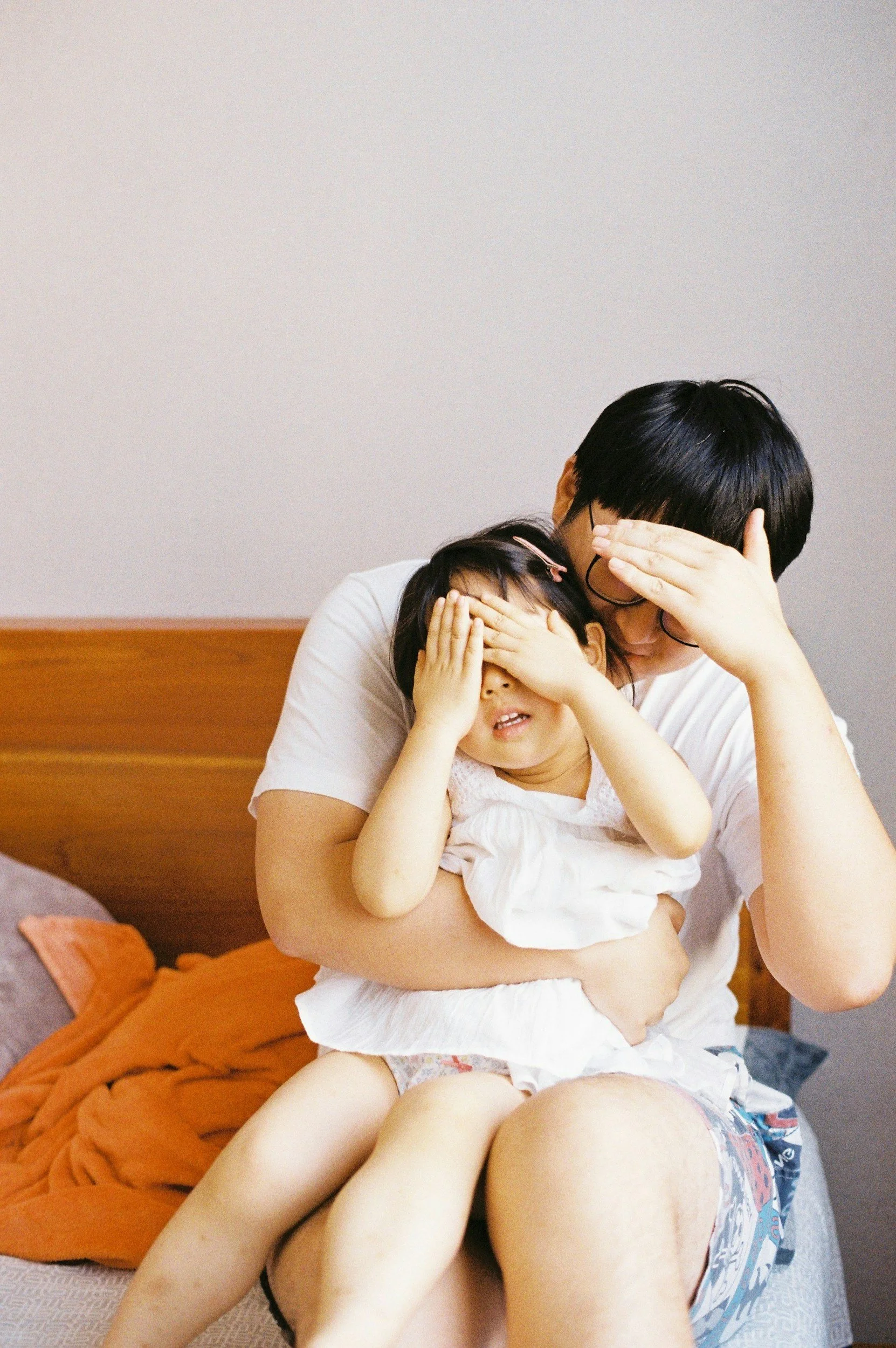 A parent sitting on a couch holding a young girl on their lap, both covering their eyes with their hands.