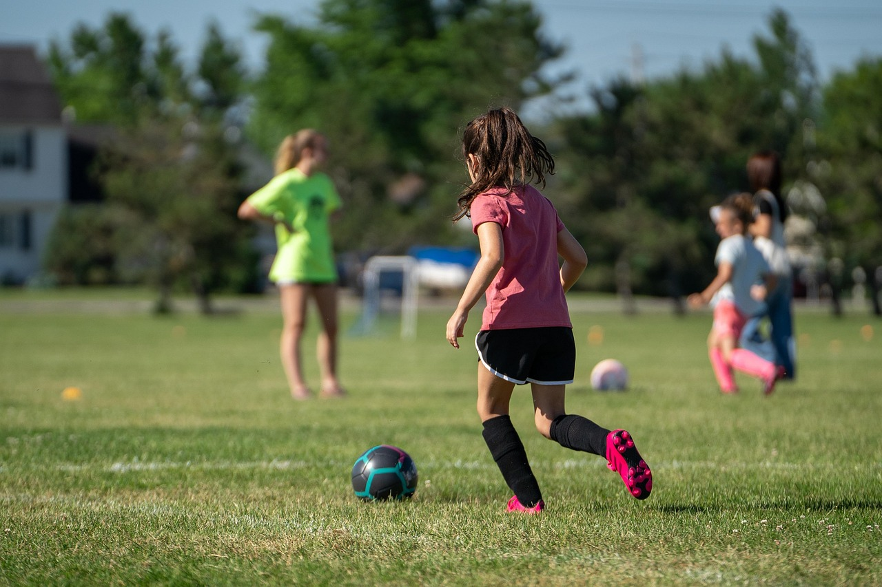 Young girl playing soccer on a grassy field, with other girls and an adult in the background, some trees, a soccer ball, and a goalpost.
