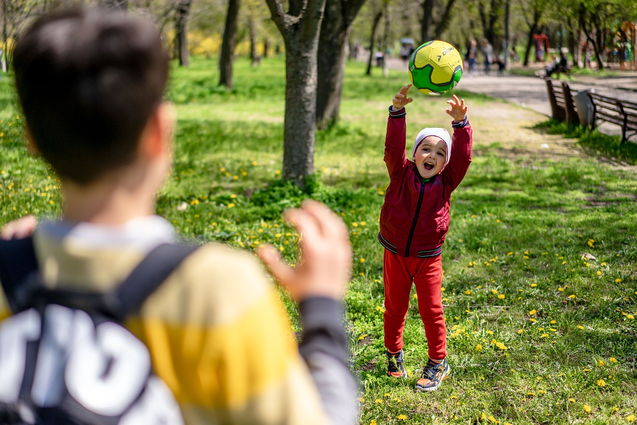 A young girl in a red jacket catching a green and yellow soccer ball in a park with trees and benches in the background.