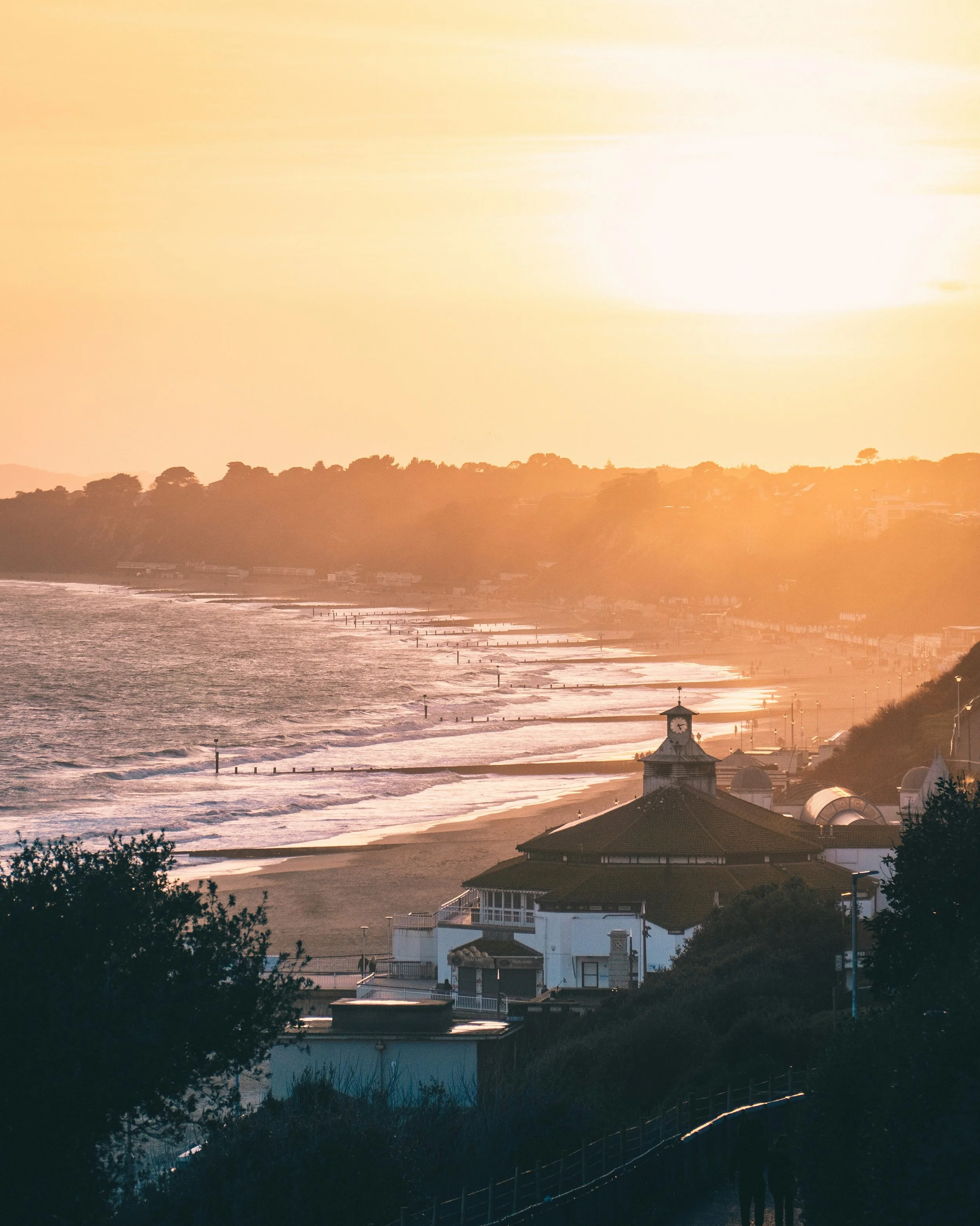 Sunset over a beach with waves crashing on the shore, silhouetted houses and trees on the hillside, and people walking along the path.